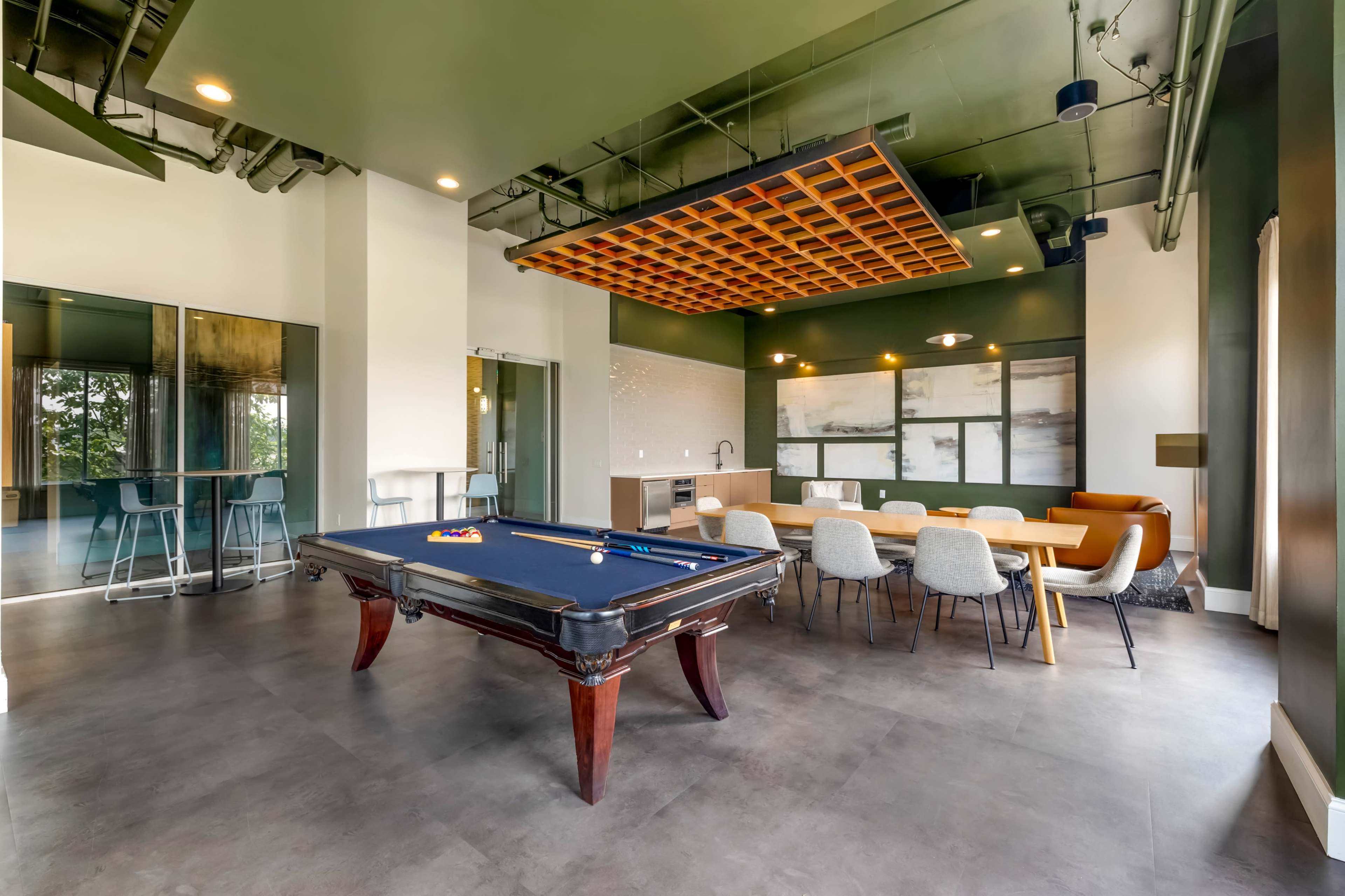 The image shows a modern game room featuring a pool table, a dining area with chairs, and a kitchenette under a decorative ceiling.