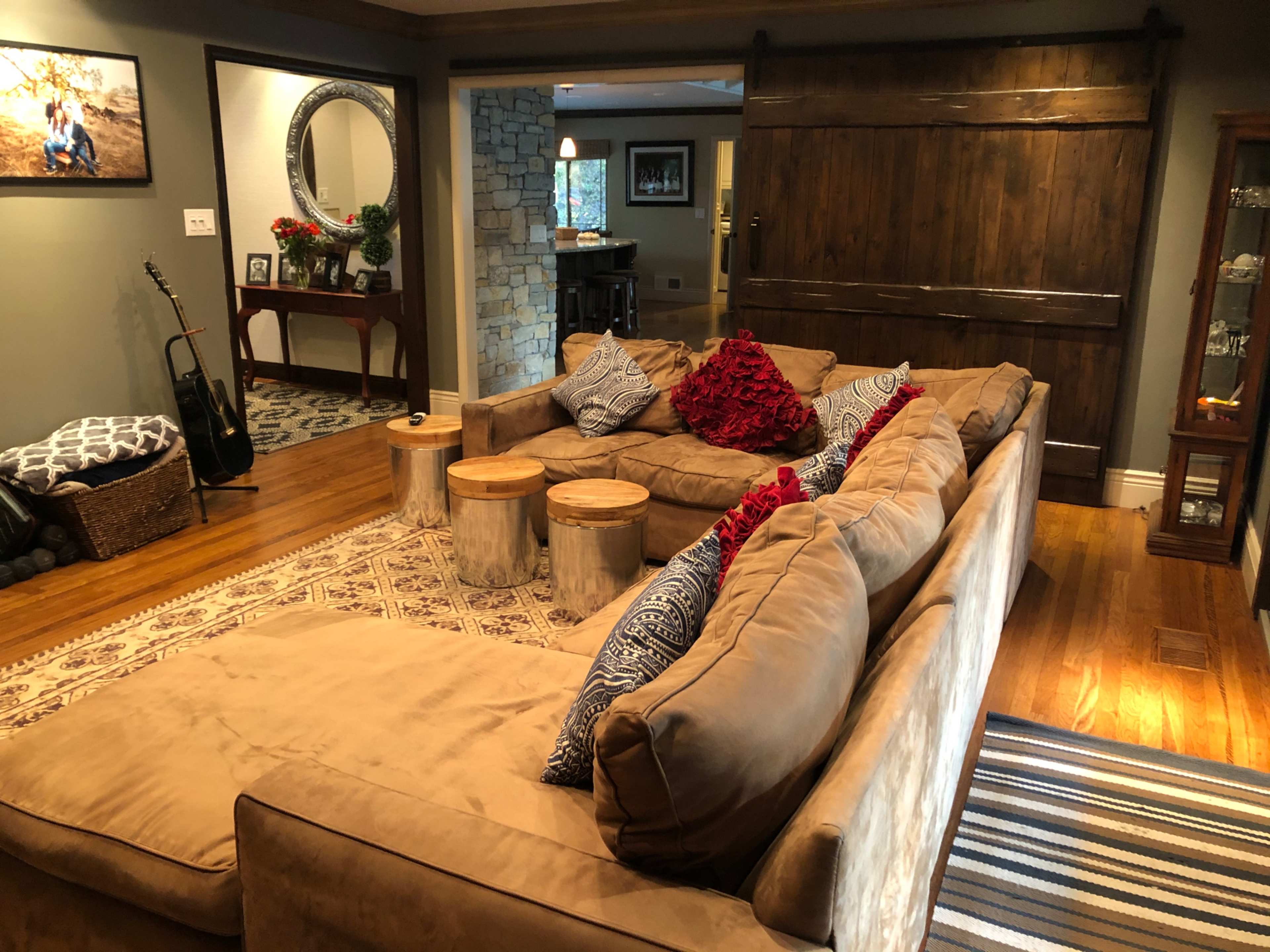A spacious living room features a large, sectional sofa with red and blue decorative pillows, wooden side tables, and a backdrop of stone walls and a large, sliding barn door.