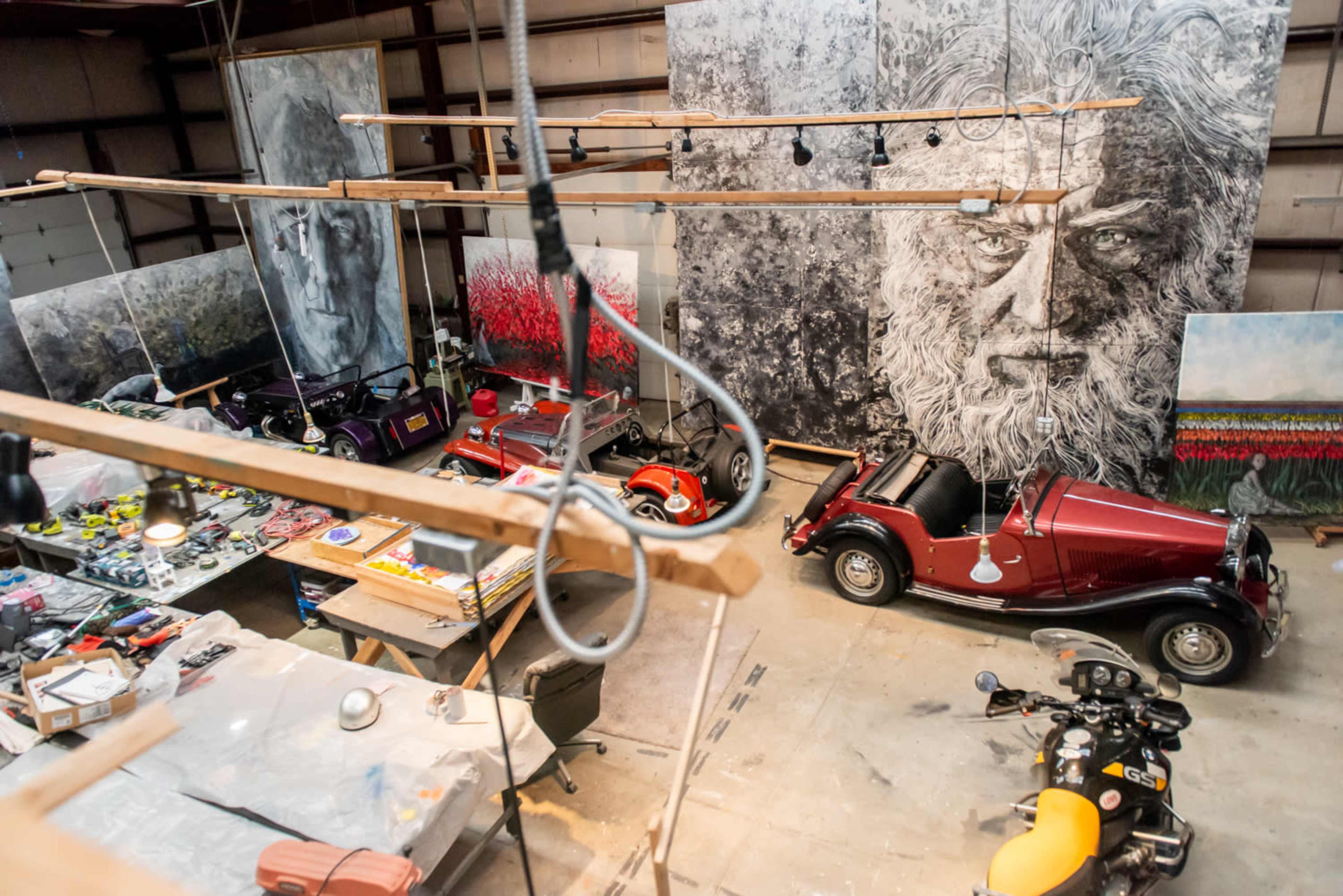 An artist's studio featuring multiple large paintings, several vintage cars, and a cluttered workspace.