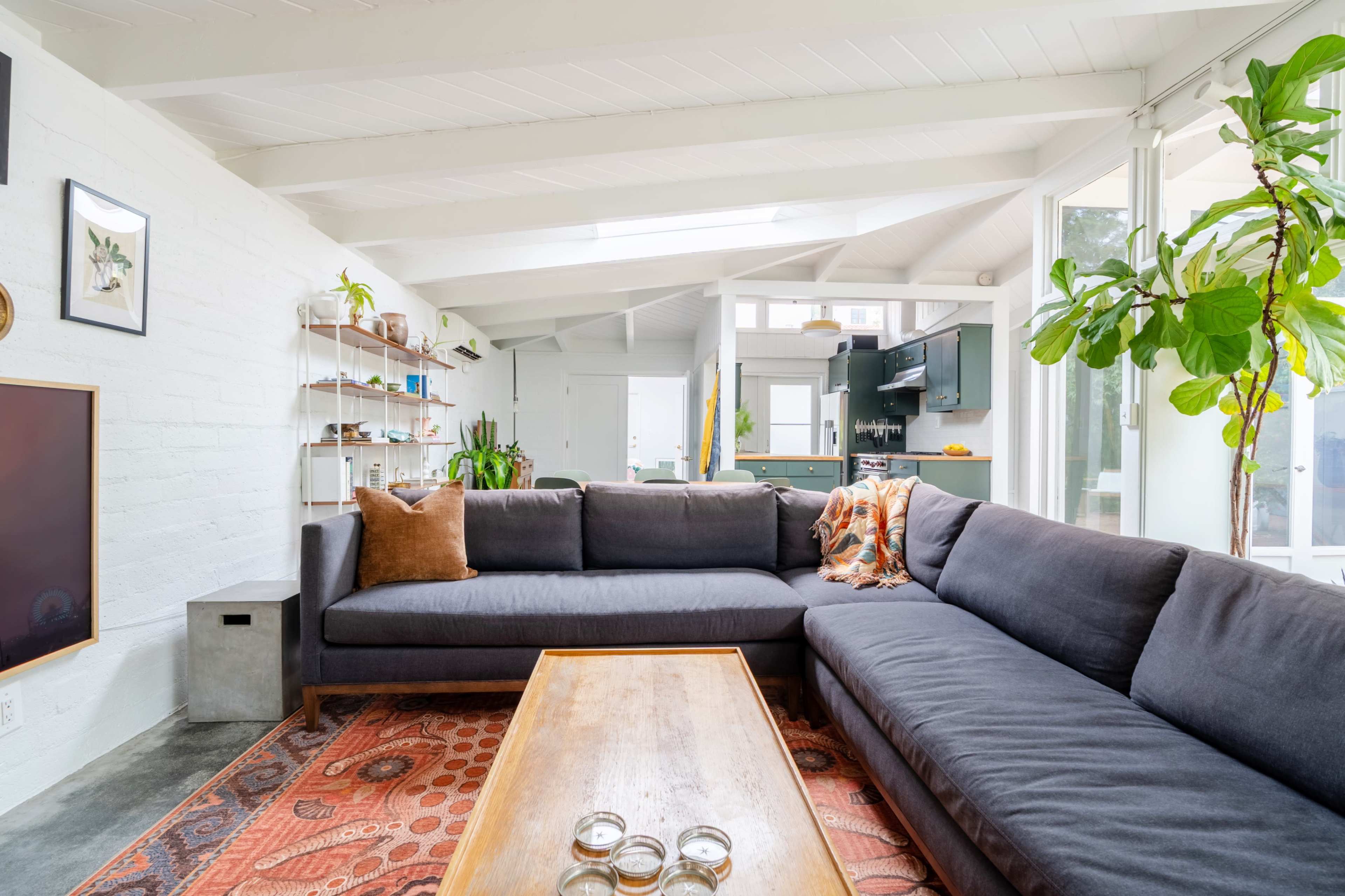 The image shows a spacious living room featuring a large sectional sofa, a wooden coffee table, and plants, with light streaming in through windows.