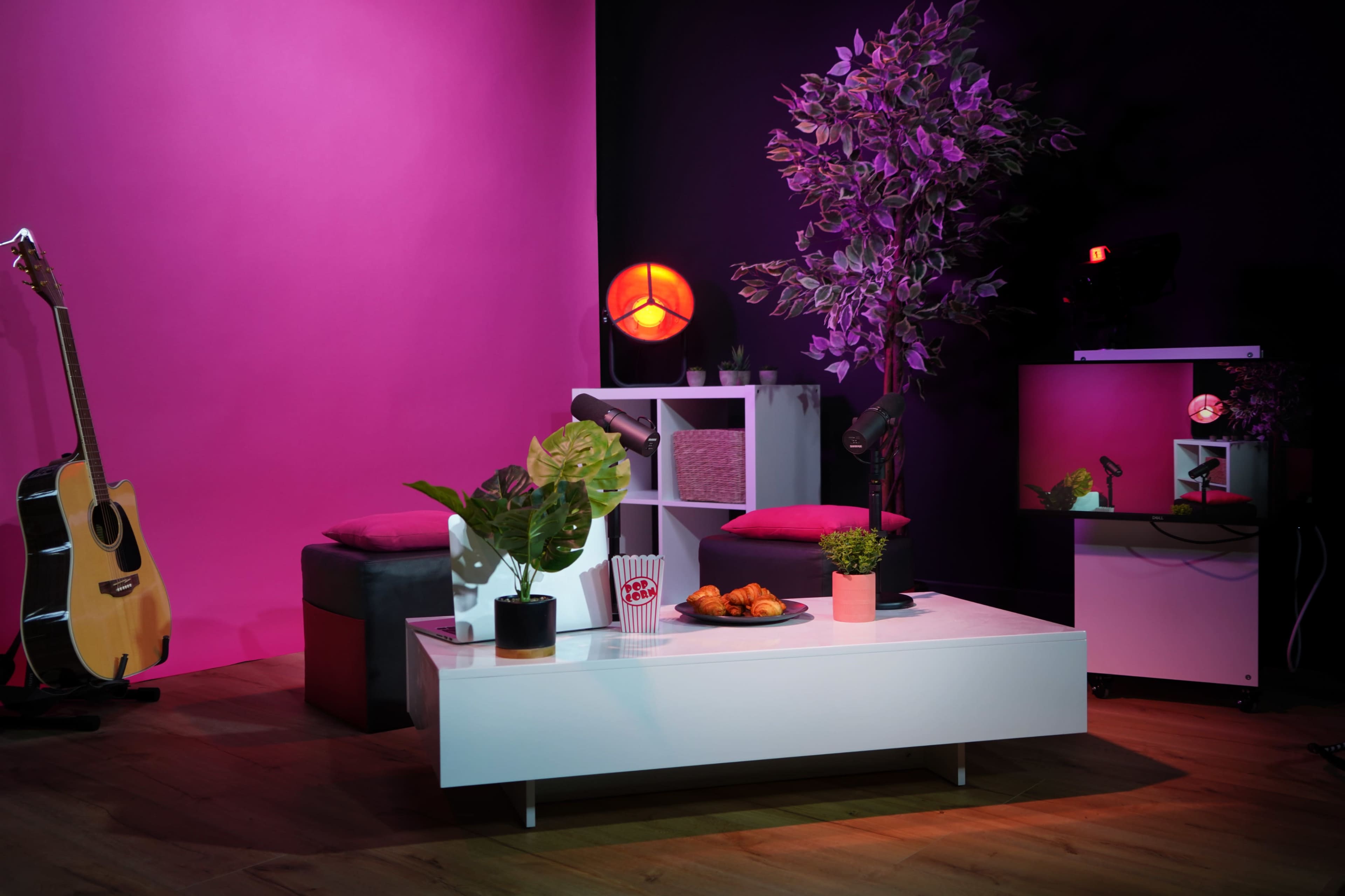 A modern living room setup features a white coffee table with snacks, plants, and a guitar beside a purple-lit backdrop.