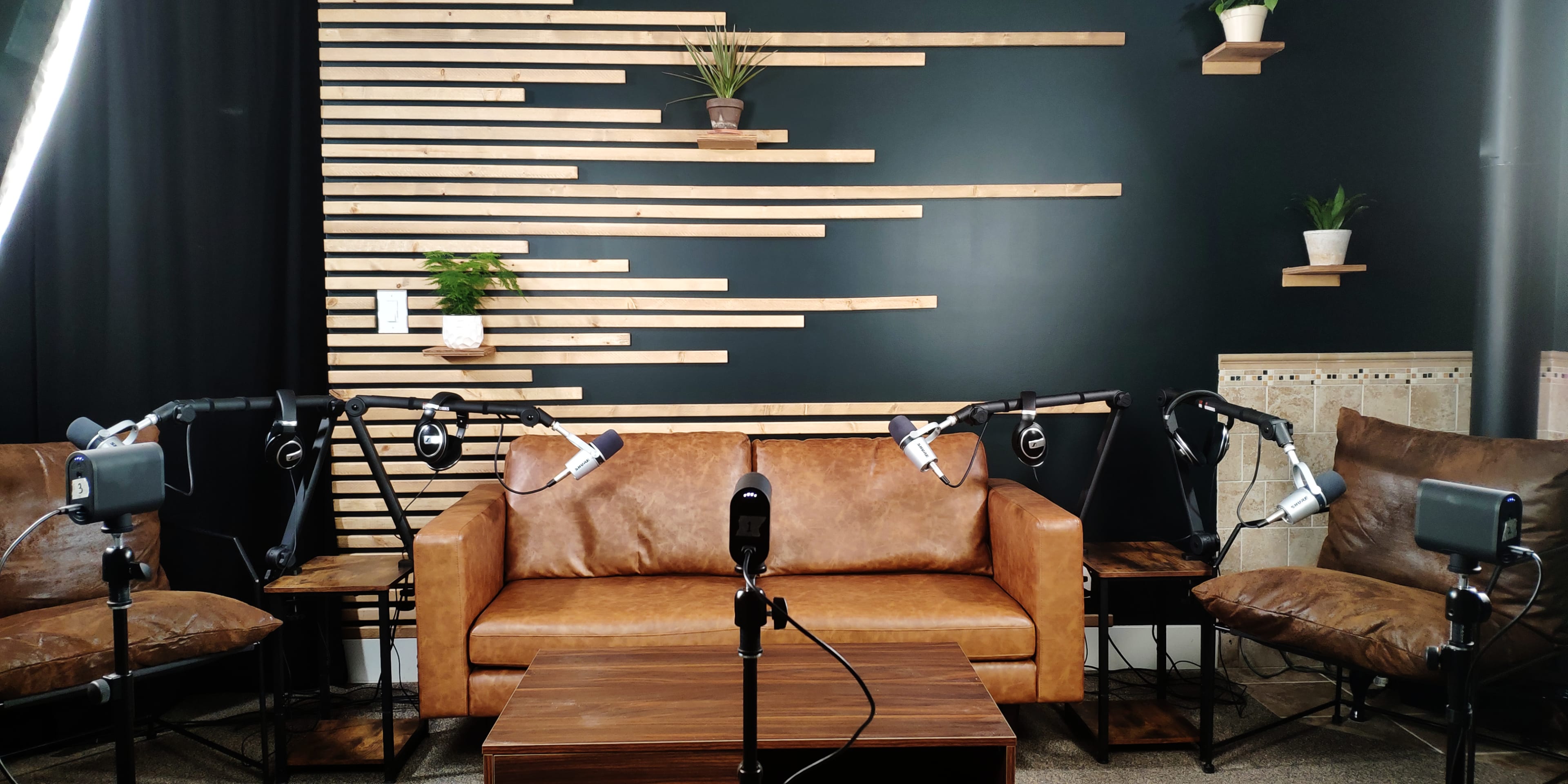 A cozy recording studio setup featuring a brown leather sofa, a wooden coffee table, and several microphone stands arranged in front of a textured wall with wooden shelves and potted plants.