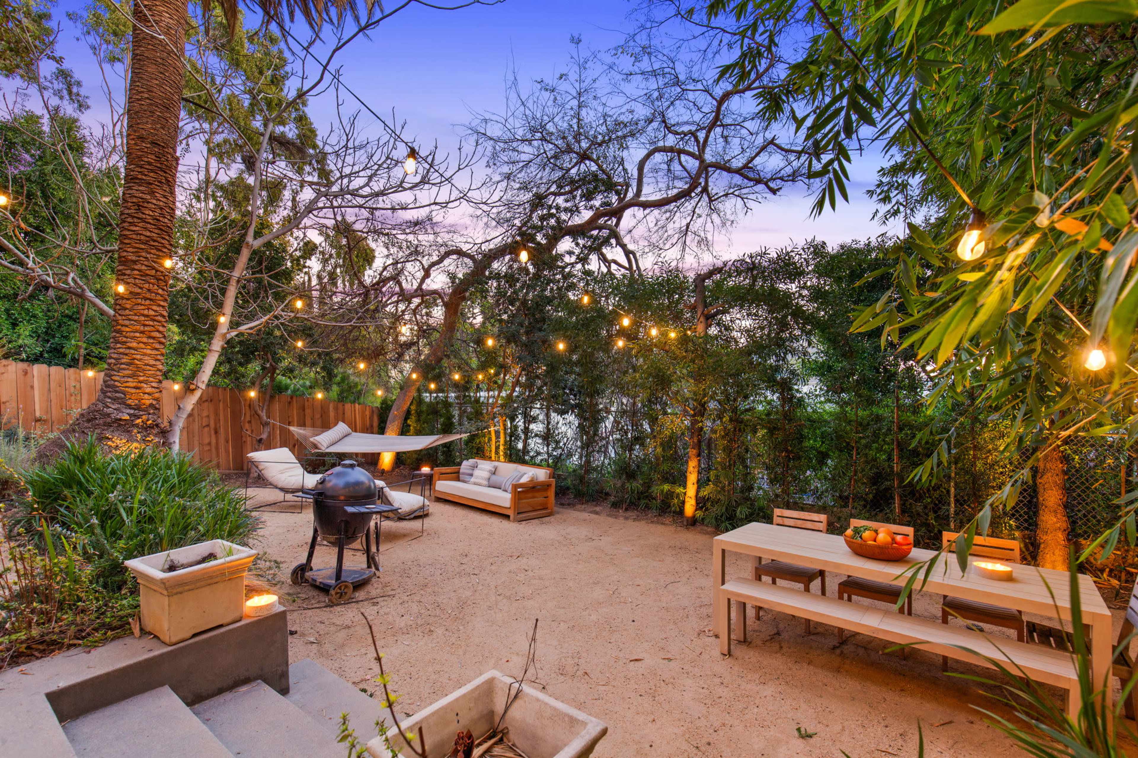 A backyard area with a dining table, seating arrangements, a barbecue grill, and string lights strung between trees.
