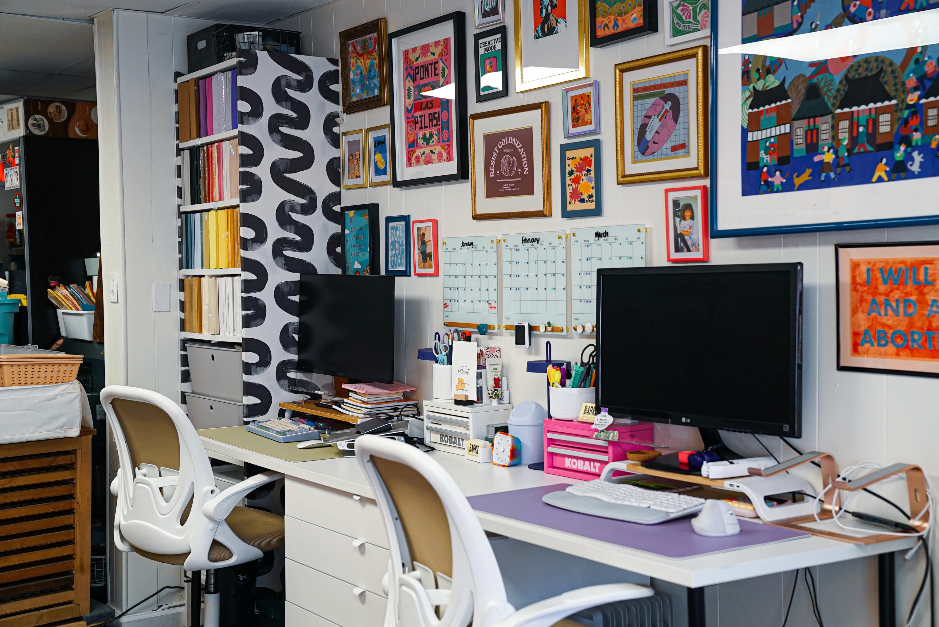 The image shows a colorful workspace featuring two computer monitors on a desk, surrounded by shelves filled with art supplies, and walls decorated with framed artwork and posters.