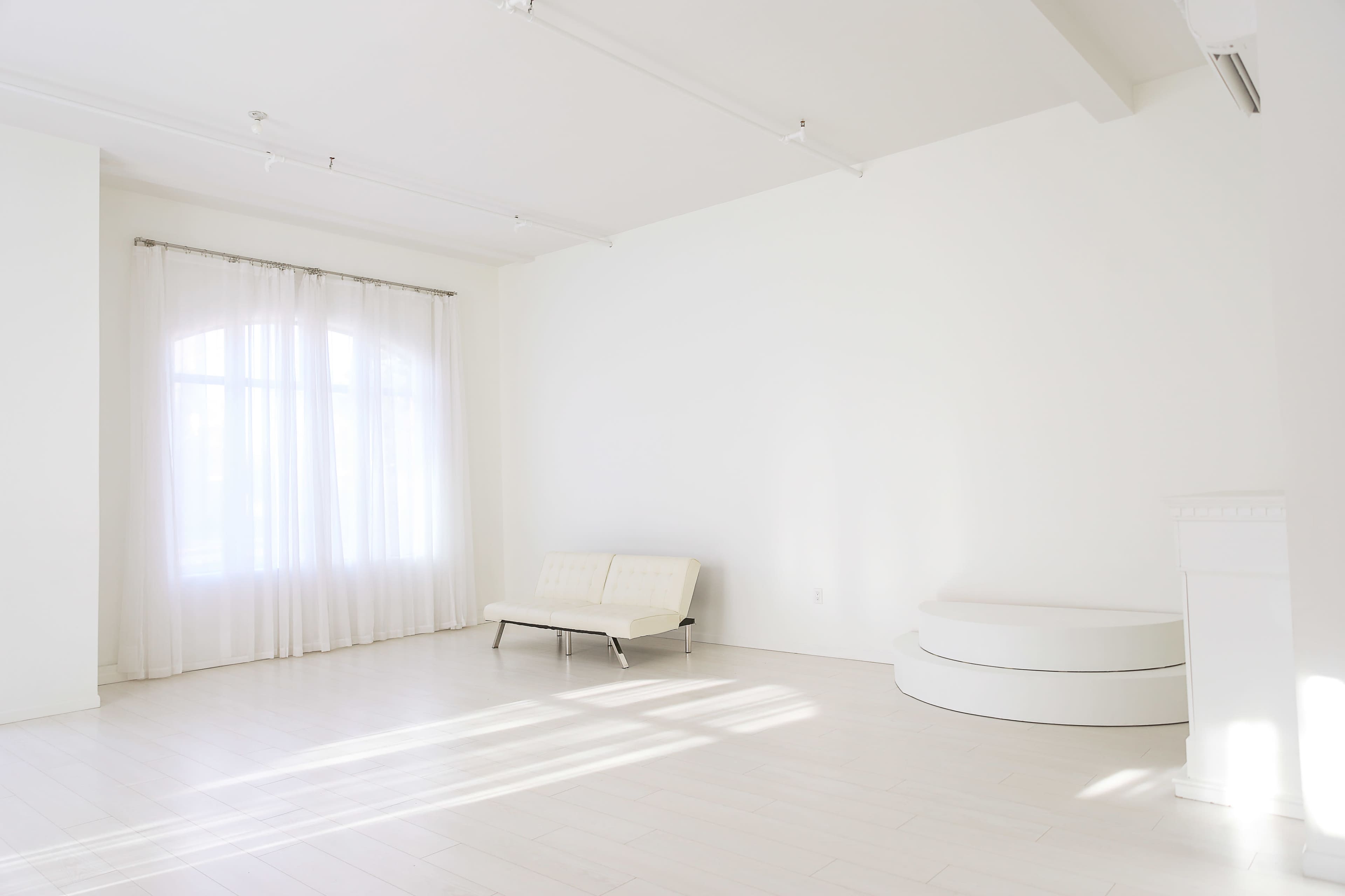 A bright, empty room features a white sofa, a curved white platform, and large windows covered with sheer curtains.
