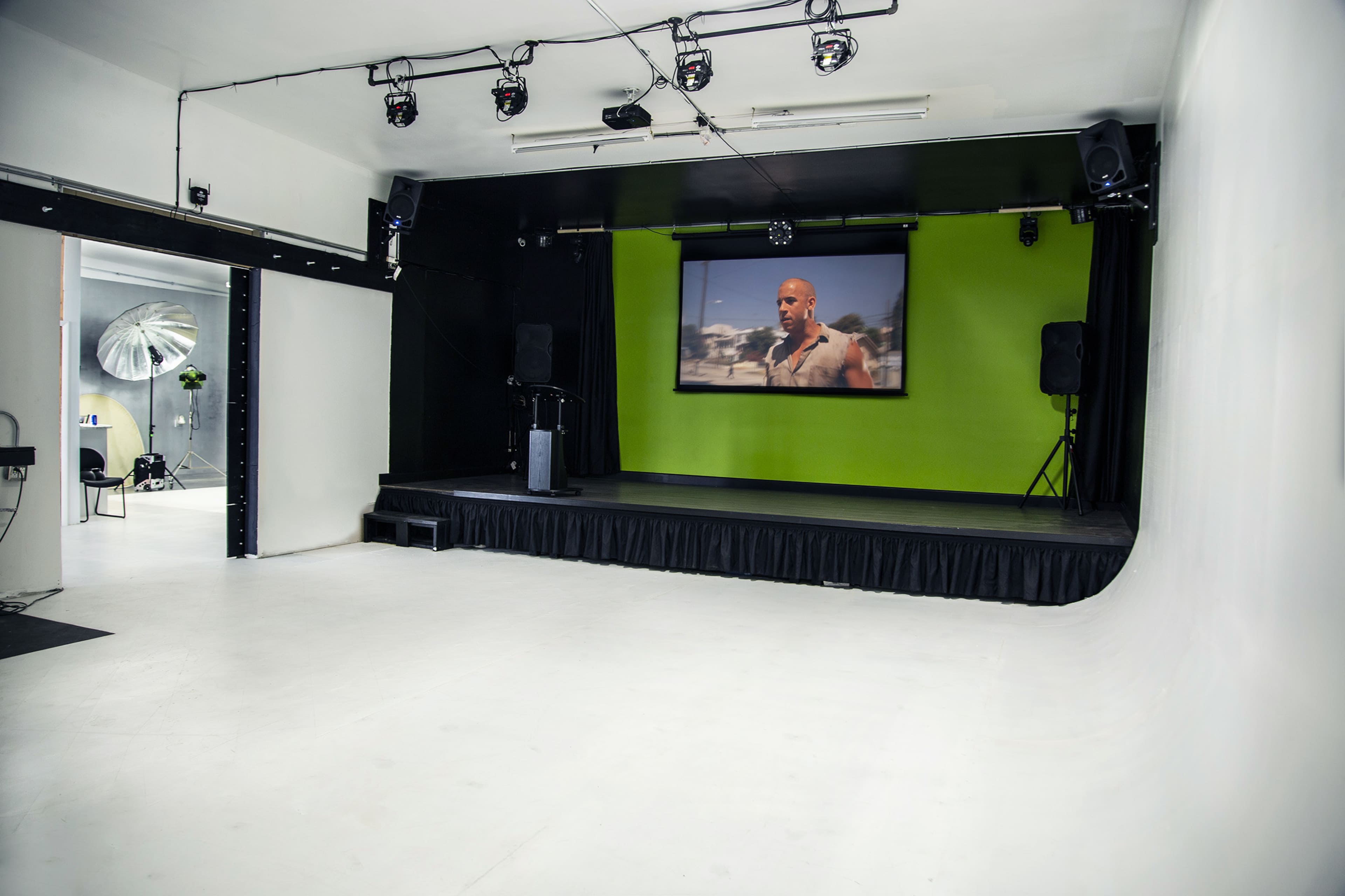 An empty stage with a green backdrop, a large screen displaying a scene, and audio equipment setup on either side.