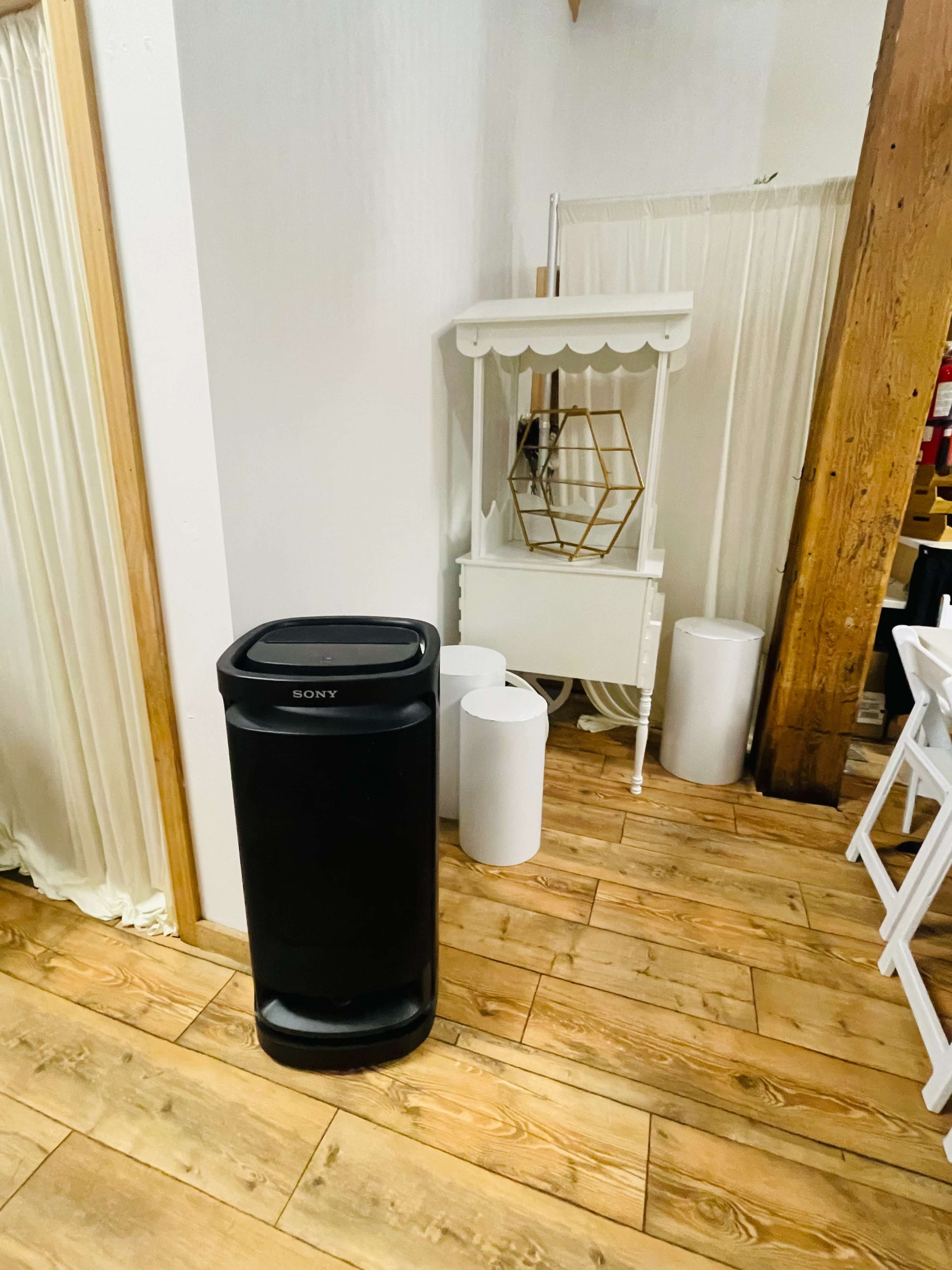 The image shows a black Sony trash can positioned next to a white decorative cabinet and a white cylindrical object in a room with wooden flooring.