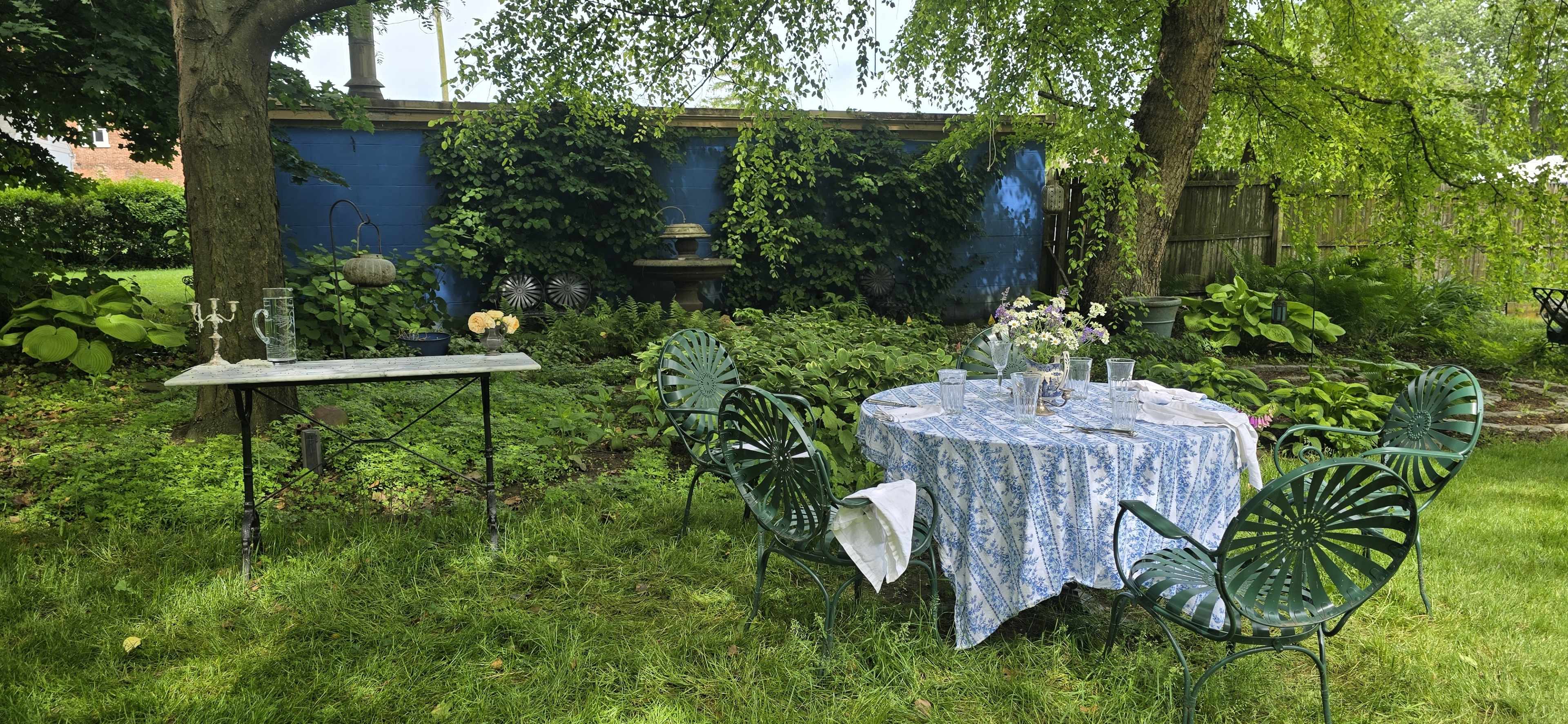 A table set with a blue and white tablecloth is surrounded by green chairs in a garden with lush foliage and a blue wall in the background.