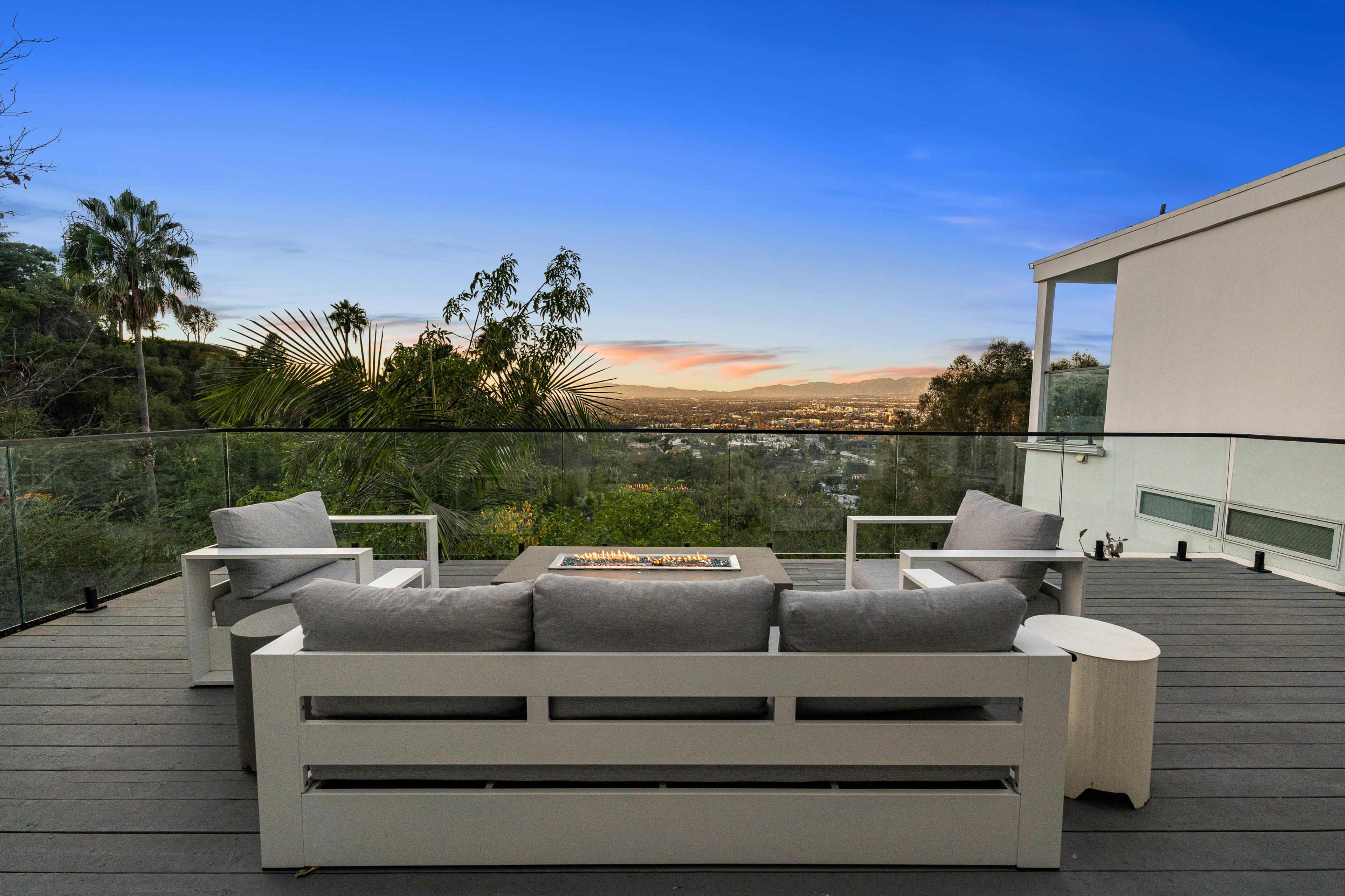 A modern outdoor seating area features gray cushions on white furniture, overlooking a scenic view at sunset from a balcony.