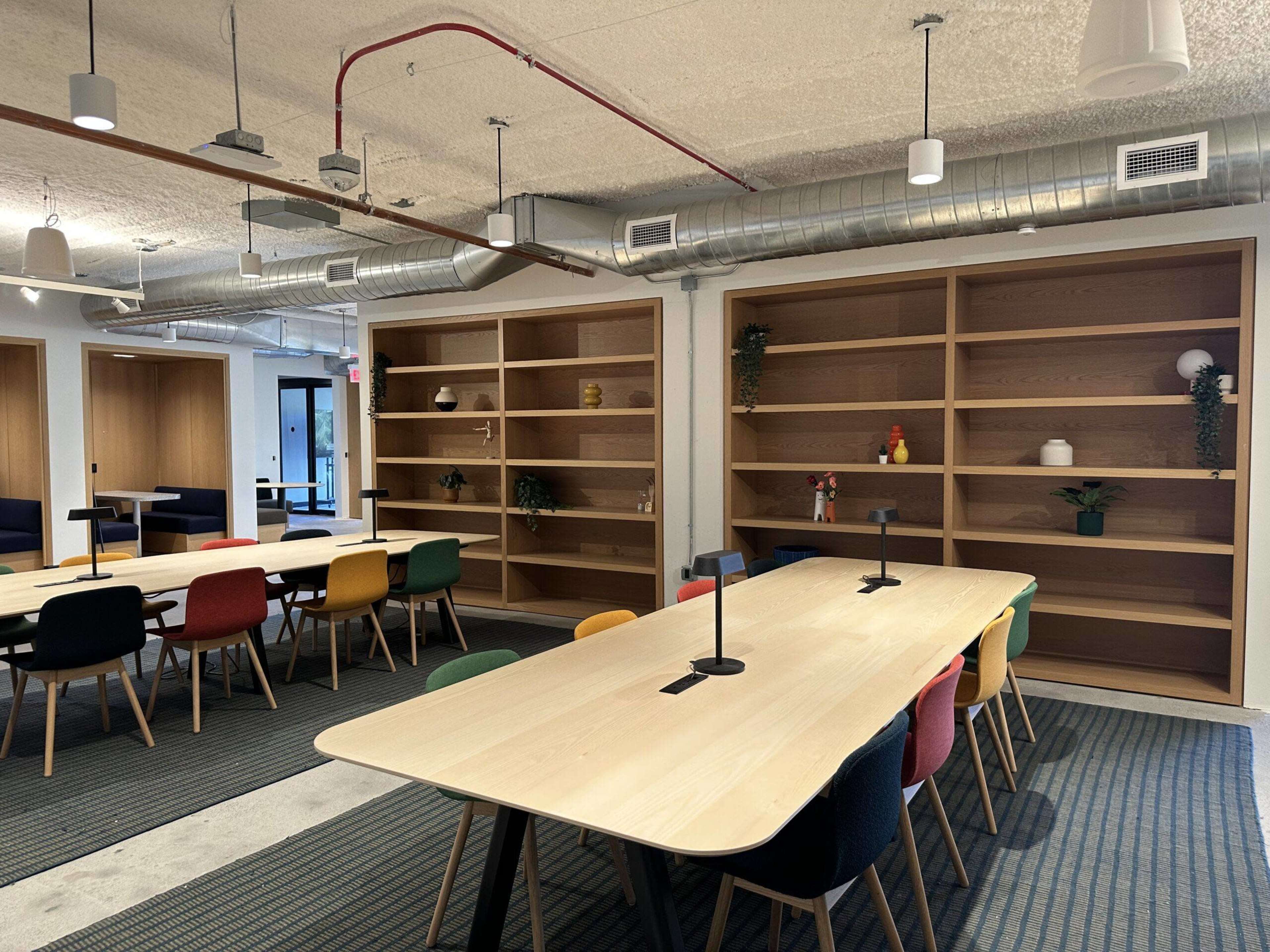 The image shows a modern office space with a long wooden table surrounded by colorful chairs and built-in wooden shelves on the walls.