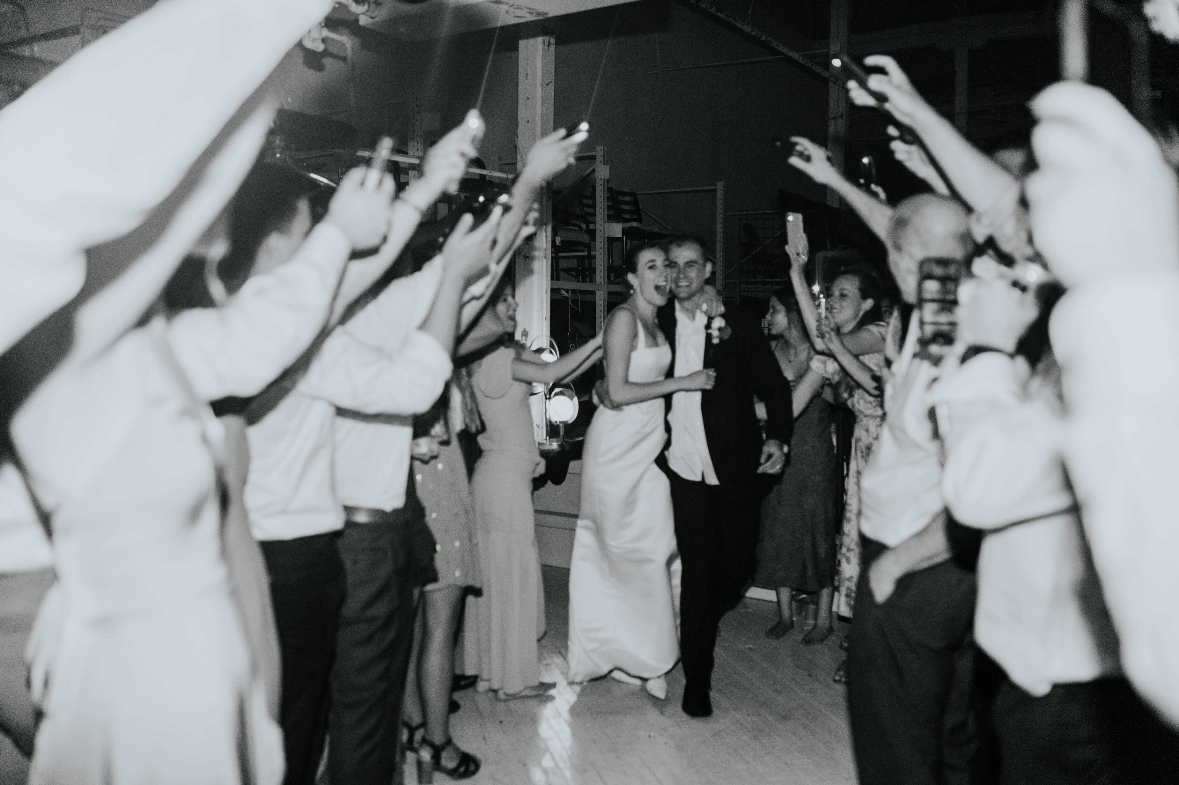 A couple exits a wedding reception while guests cheer and wave their phones.