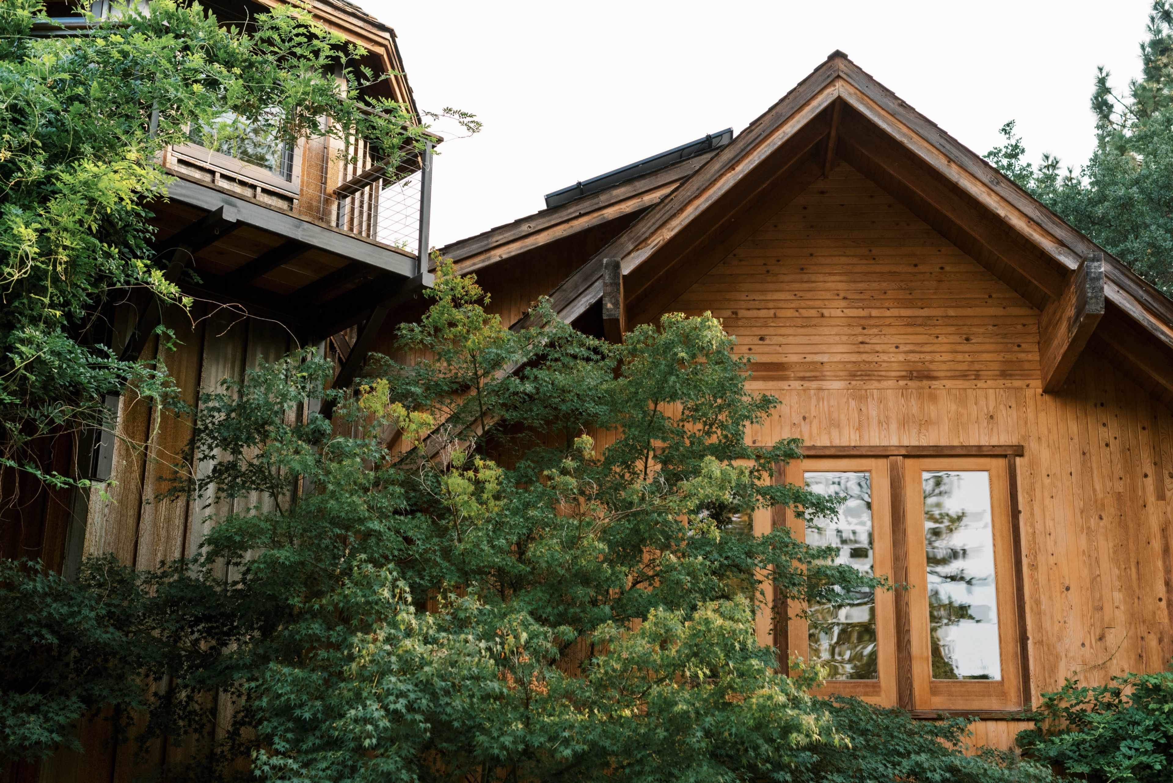 The image shows a wooden house surrounded by greenery, with large windows and a balcony on the upper level.