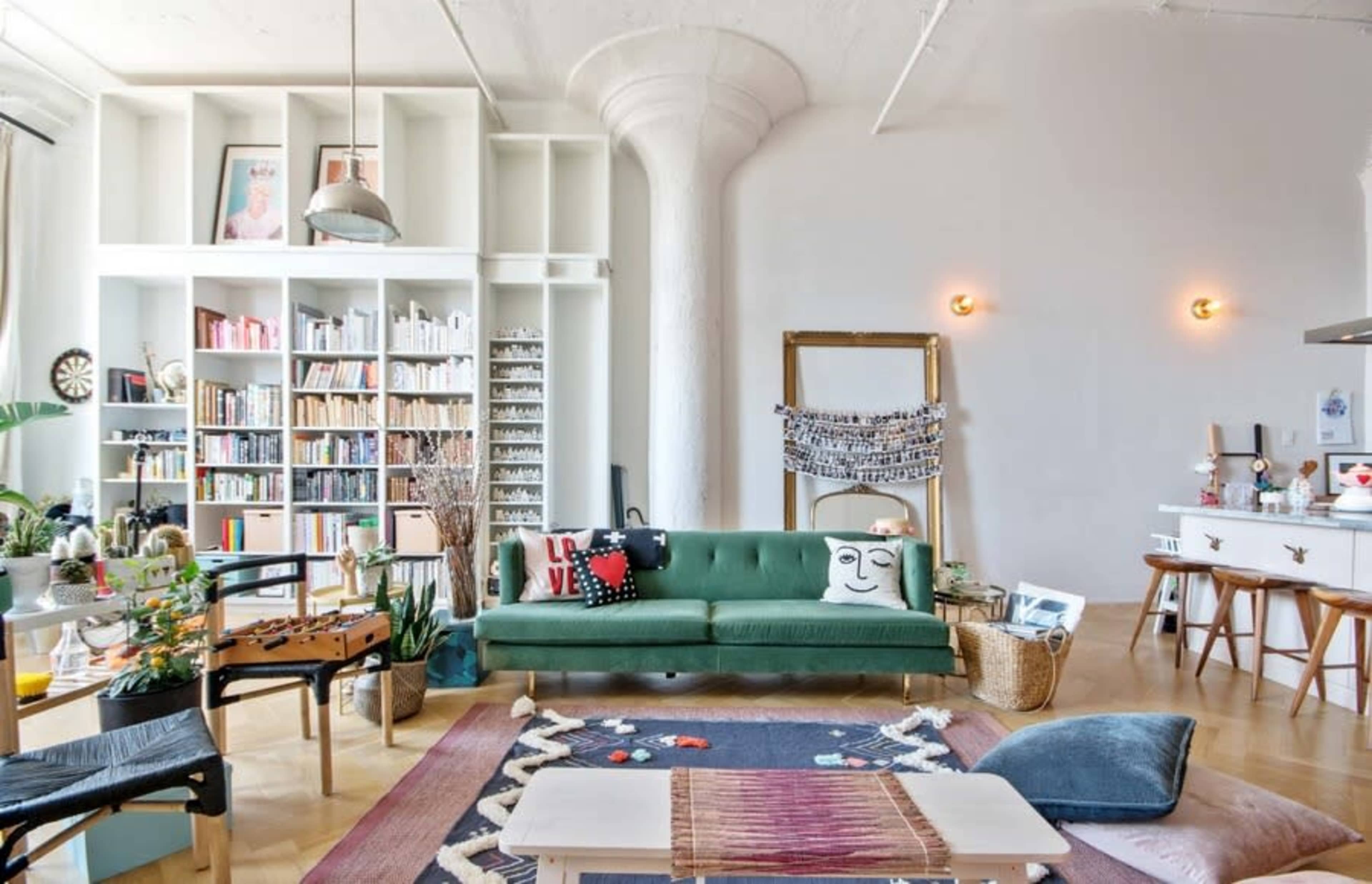 A spacious living room features a green sofa, a large bookshelf filled with books, and a mirror, with a cozy rug and various decorative items throughout the space.