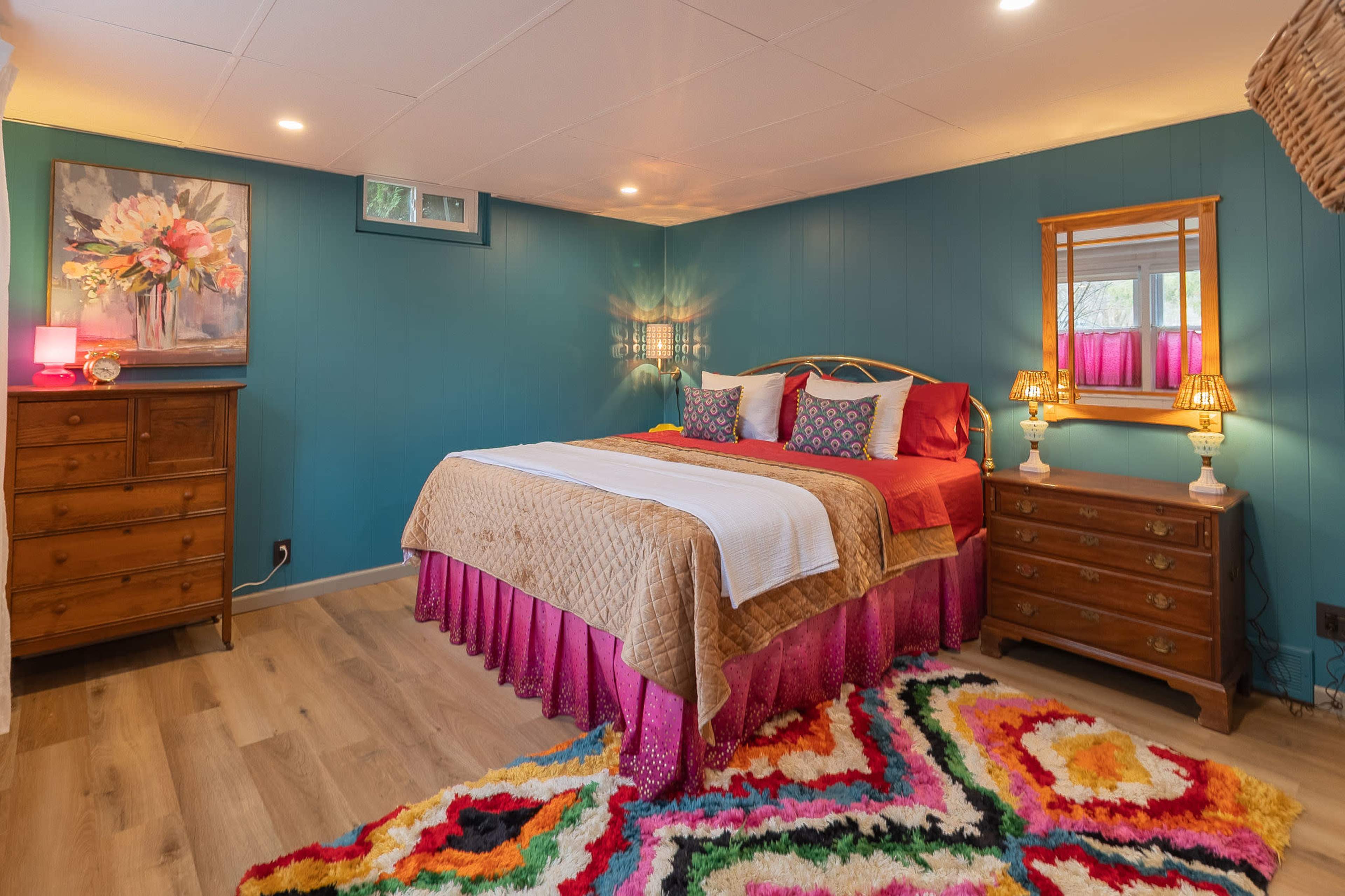 The image shows a brightly colored bedroom featuring a bed with decorative pillows, a colorful rug, wooden dressers, and wall art.