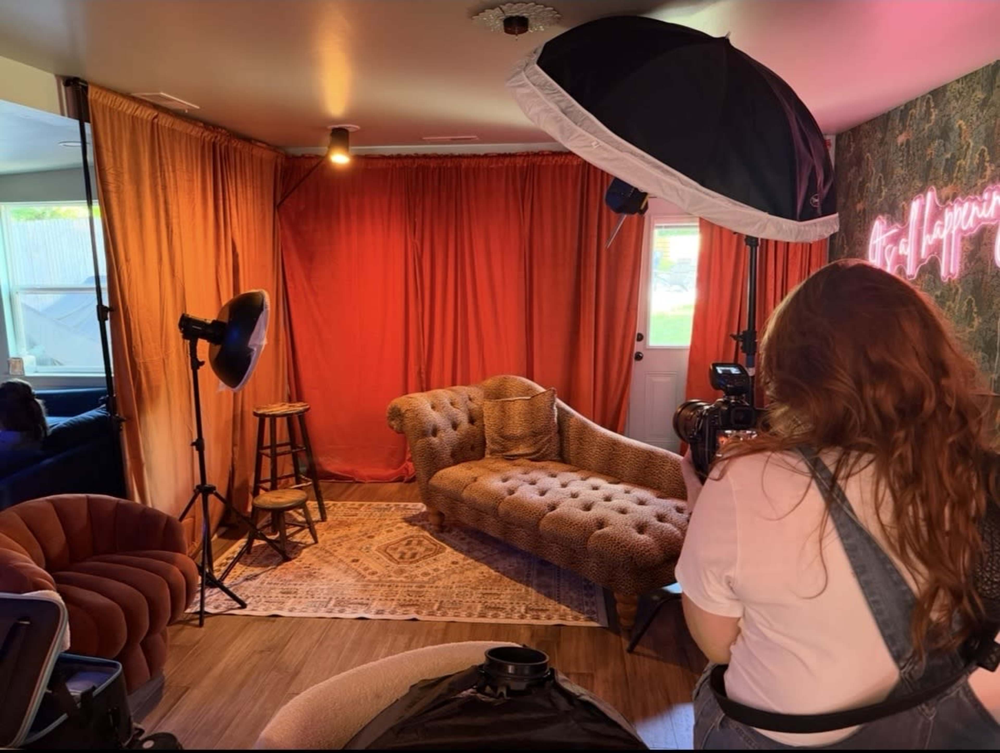 A person is setting up a photography session in a brightly lit room featuring a vintage-style couch, assorted lighting equipment, and colorful drapes.