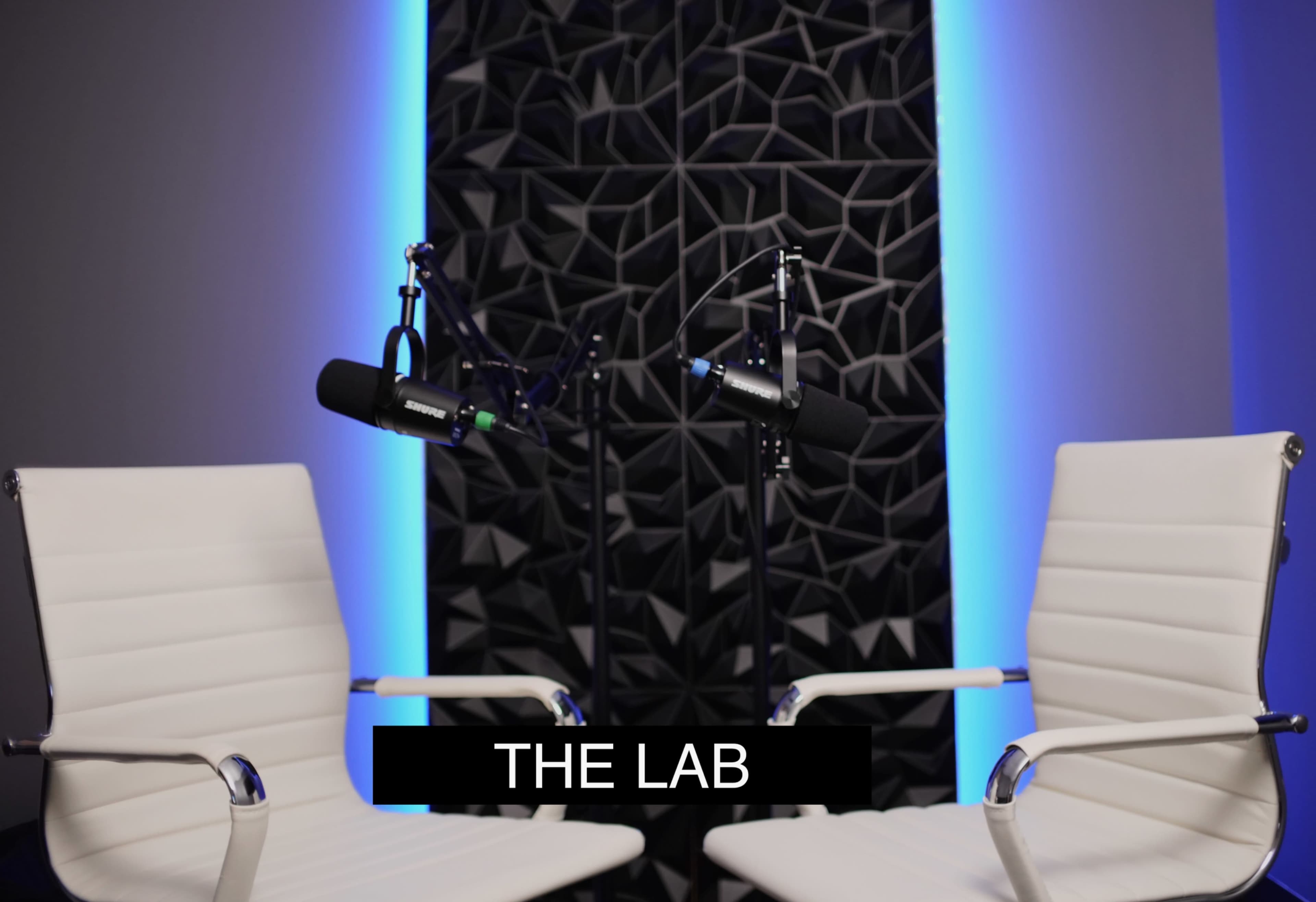 The image shows two white chairs facing each other in a modern studio setting with microphones mounted on stands and a textured wall illuminated by blue lighting.