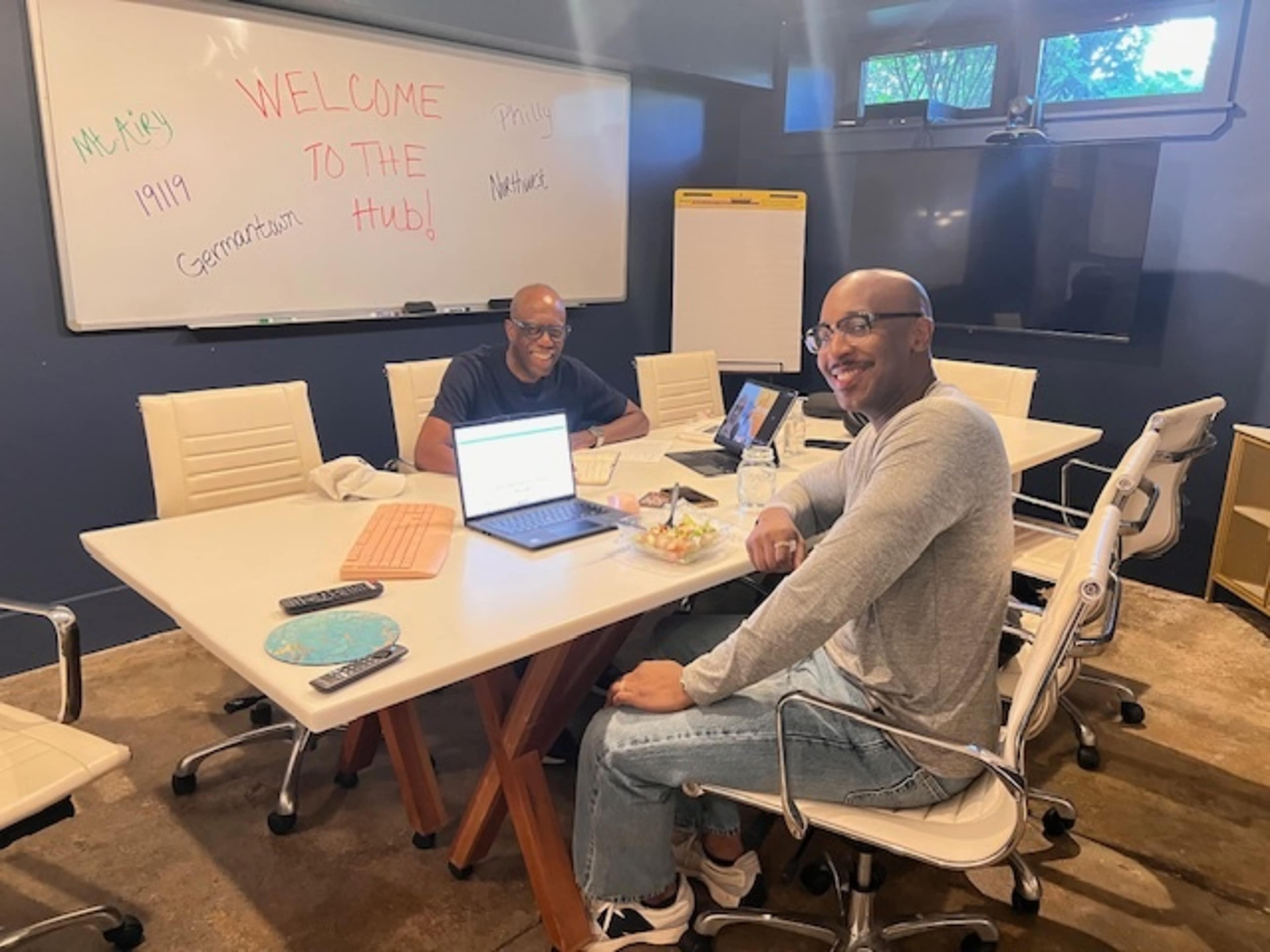 Two men are seated at a conference table in a modern meeting room with a whiteboard that says "WELCOME TO THE HUB."