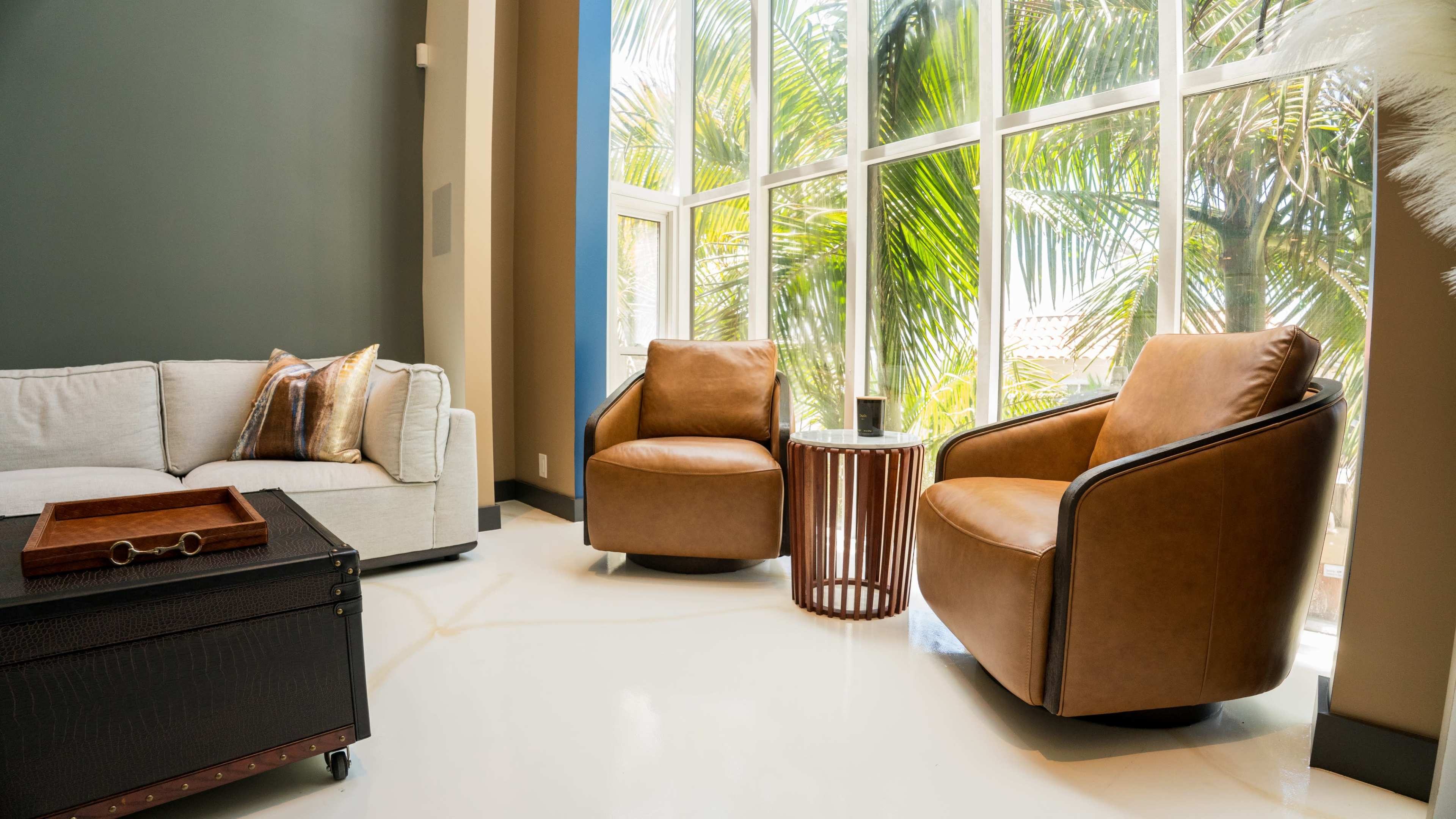 The scene features a modern living area with two brown armchairs, a small wooden side table, and a large window showcasing palm trees outside.