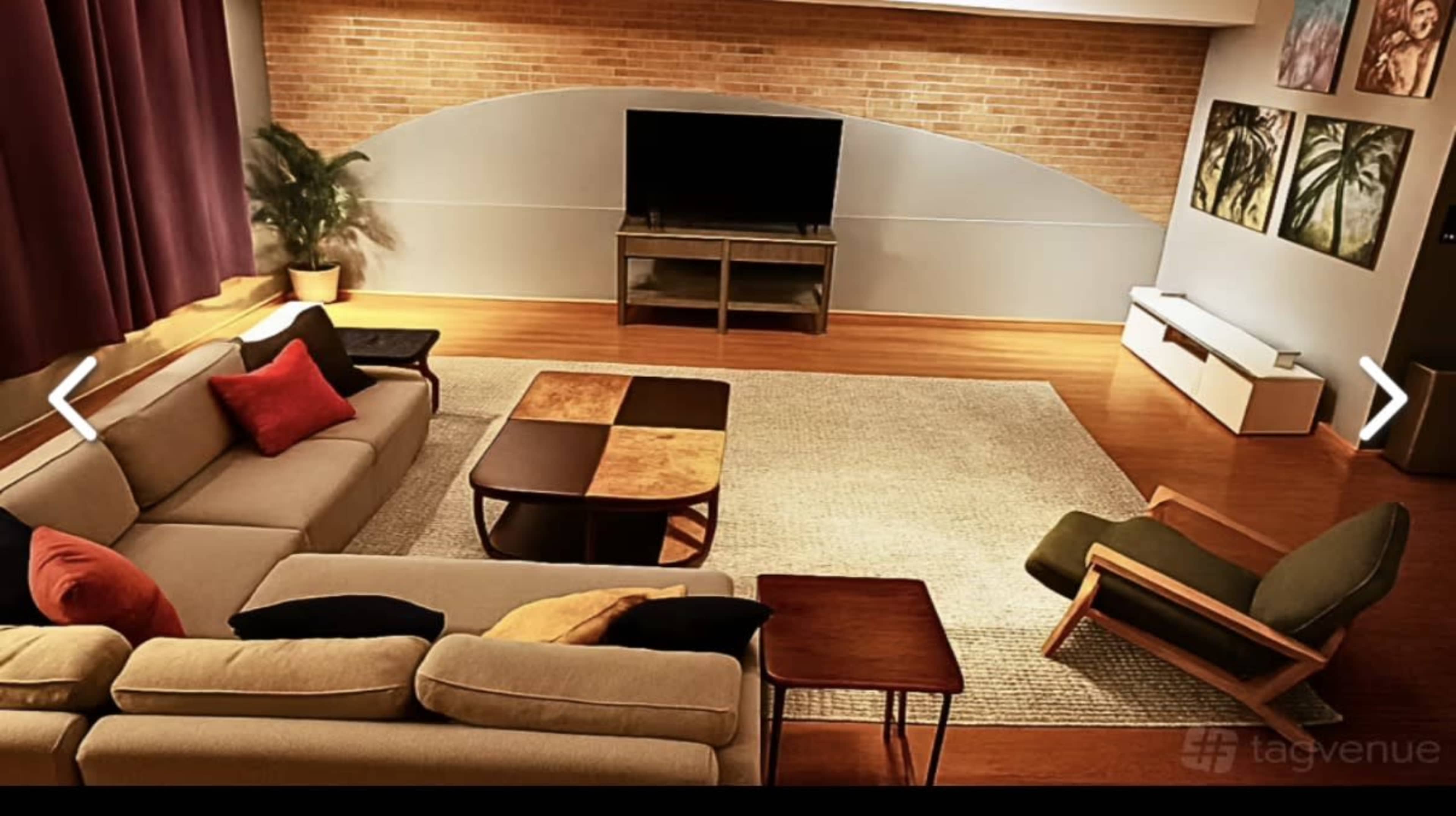 The image shows a spacious living room with a brick wall, a television on a glass cabinet, and a mix of modern furniture including a couch, coffee table, and an armchair.