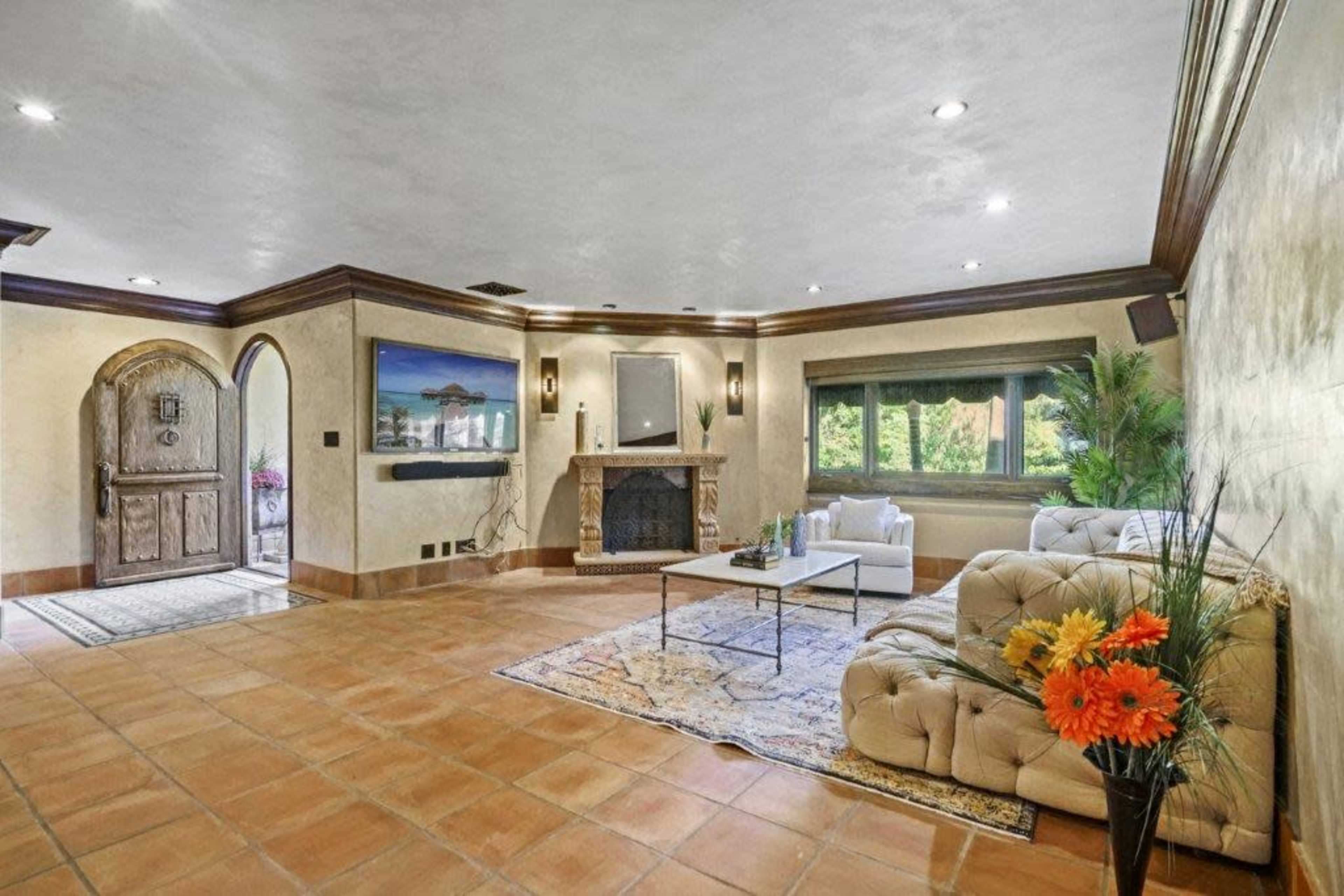 The image shows a spacious living room featuring a beige couch, a coffee table, and a fireplace, with a decorative entrance door and large windows overlooking greenery.