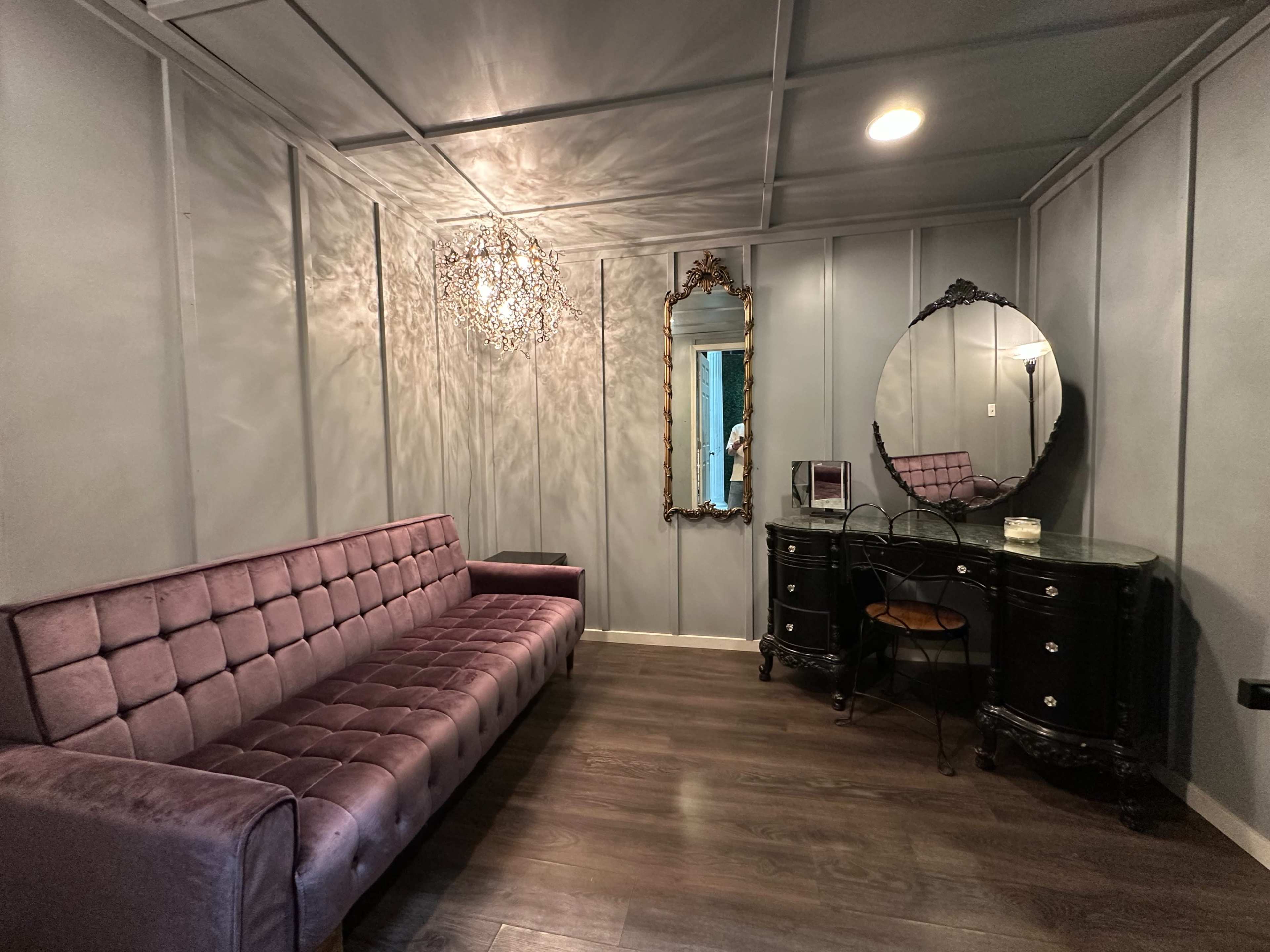 The image shows a small room with a purple velvet sofa, a decorative mirror, and a black vanity with a chair against grey paneled walls.