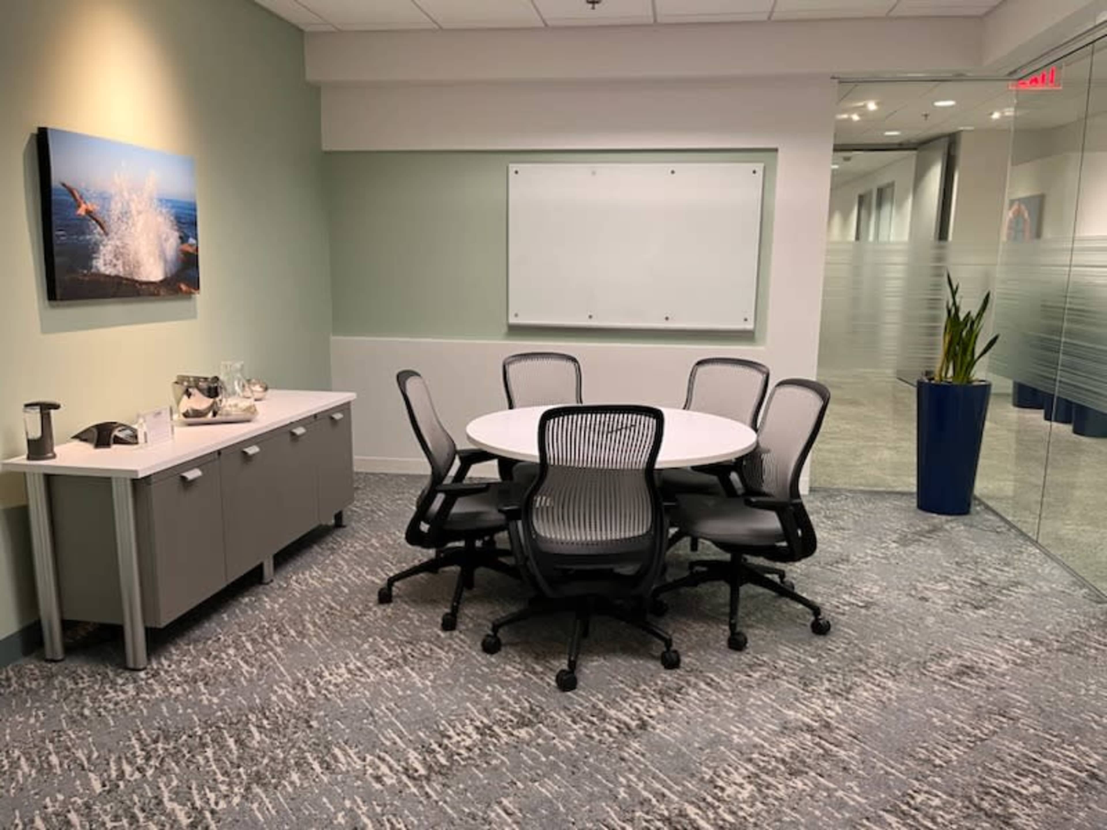 A small meeting room with a round table surrounded by five chairs, a side cabinet with a beverage setup, and a whiteboard on the wall.