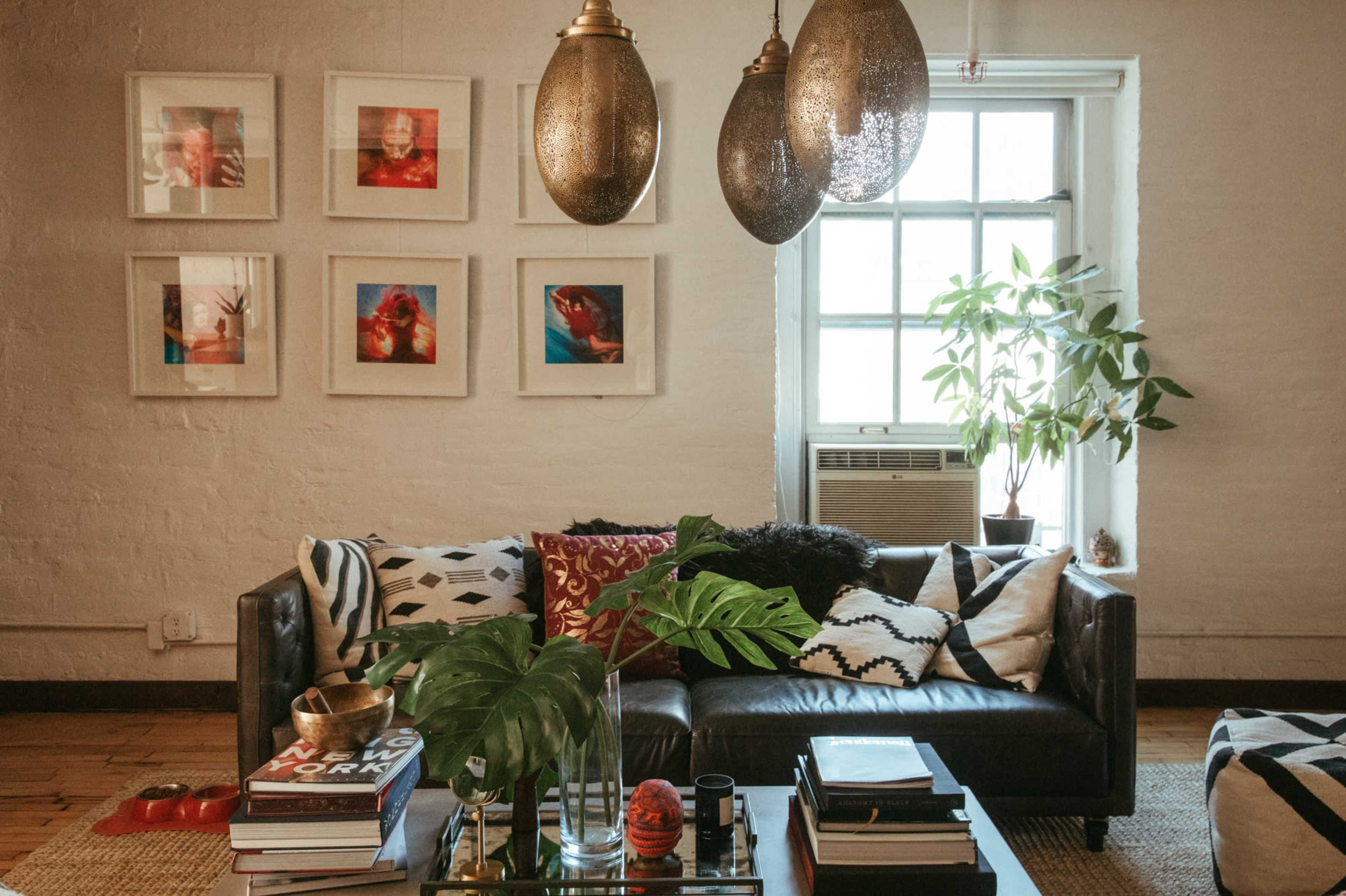 A cozy living room with a dark leather sofa, decorative pillows, a coffee table stacked with books, framed art on the wall, and a potted plant beside a window.