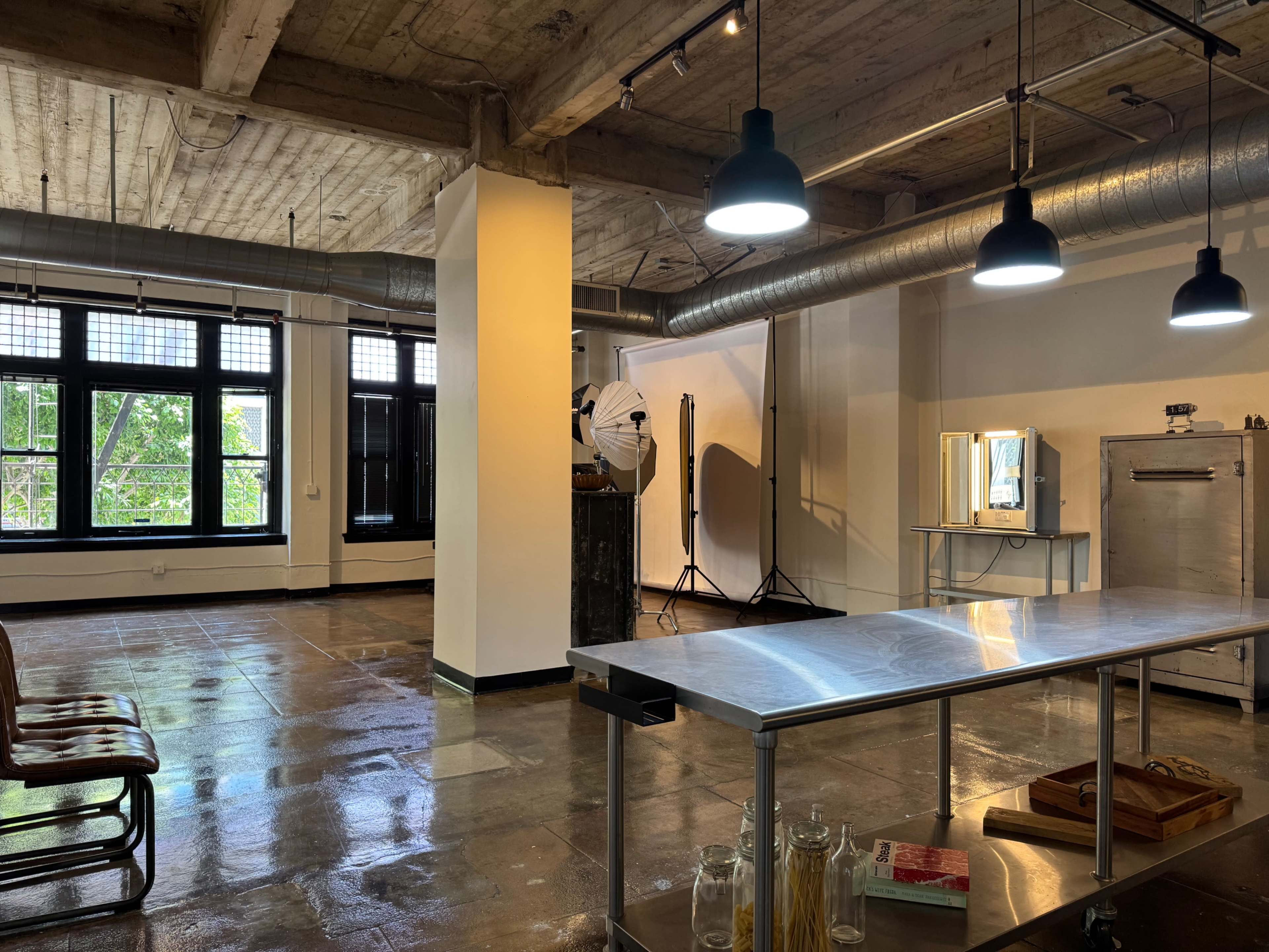 An industrial-style studio features polished concrete floors, large windows, and minimalist furnishings, including a stainless steel table and photography equipment.