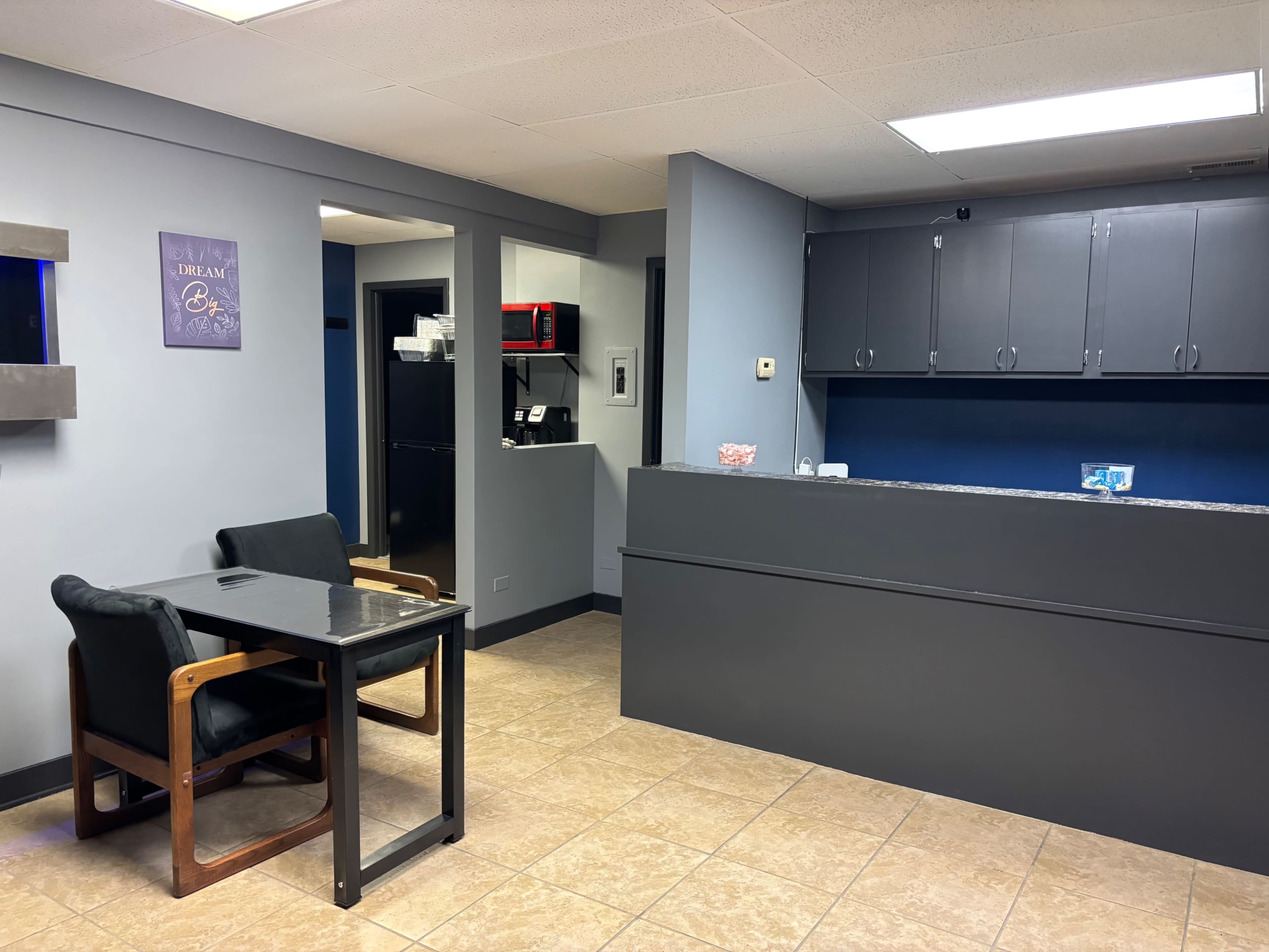 The image shows a well-lit reception area with a gray front desk, a small table with two chairs, and a kitchenette in the background.