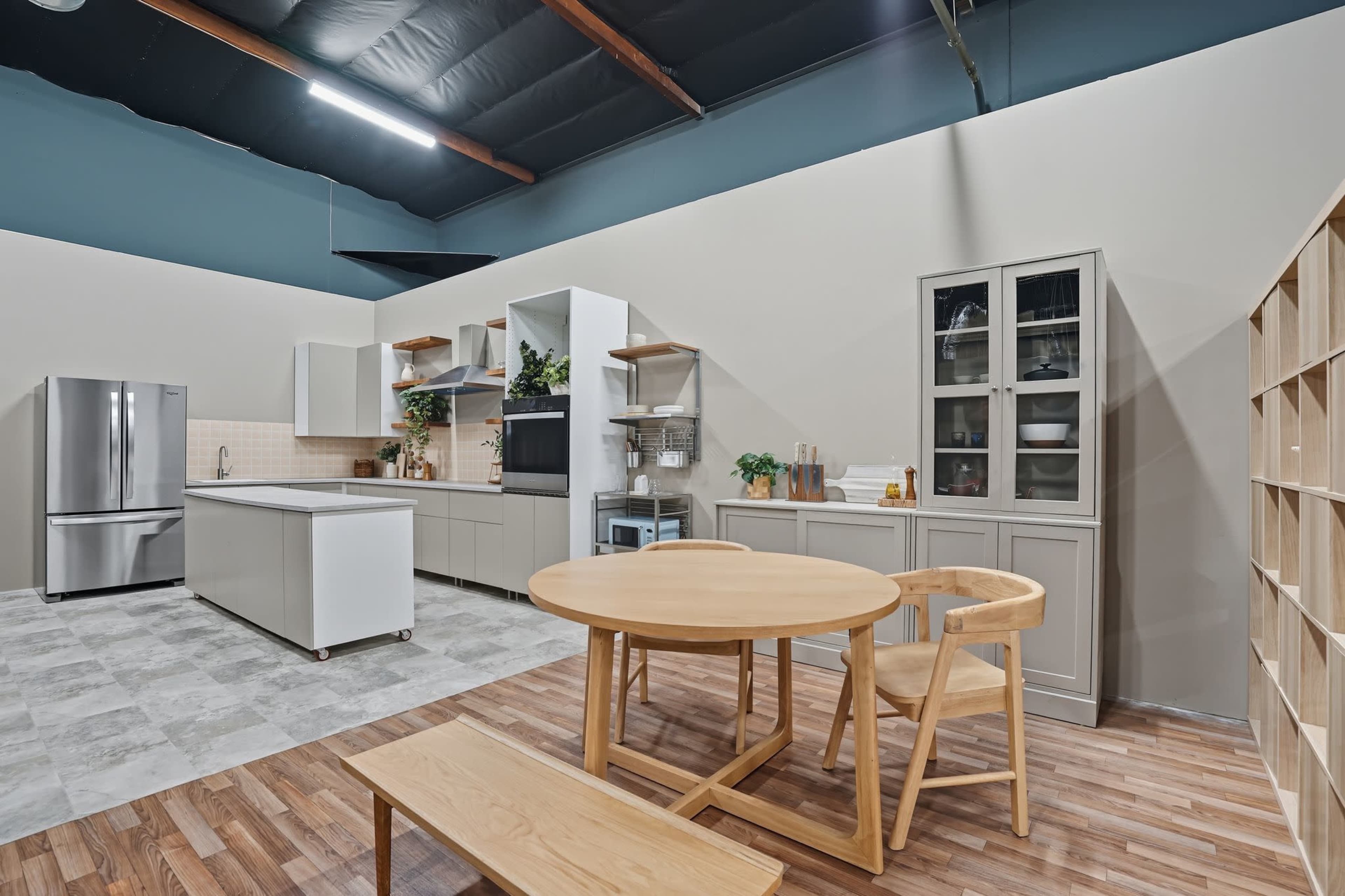 A modern kitchen features a central island, stainless steel appliances, wooden flooring, and a circular dining table paired with light-colored chairs.