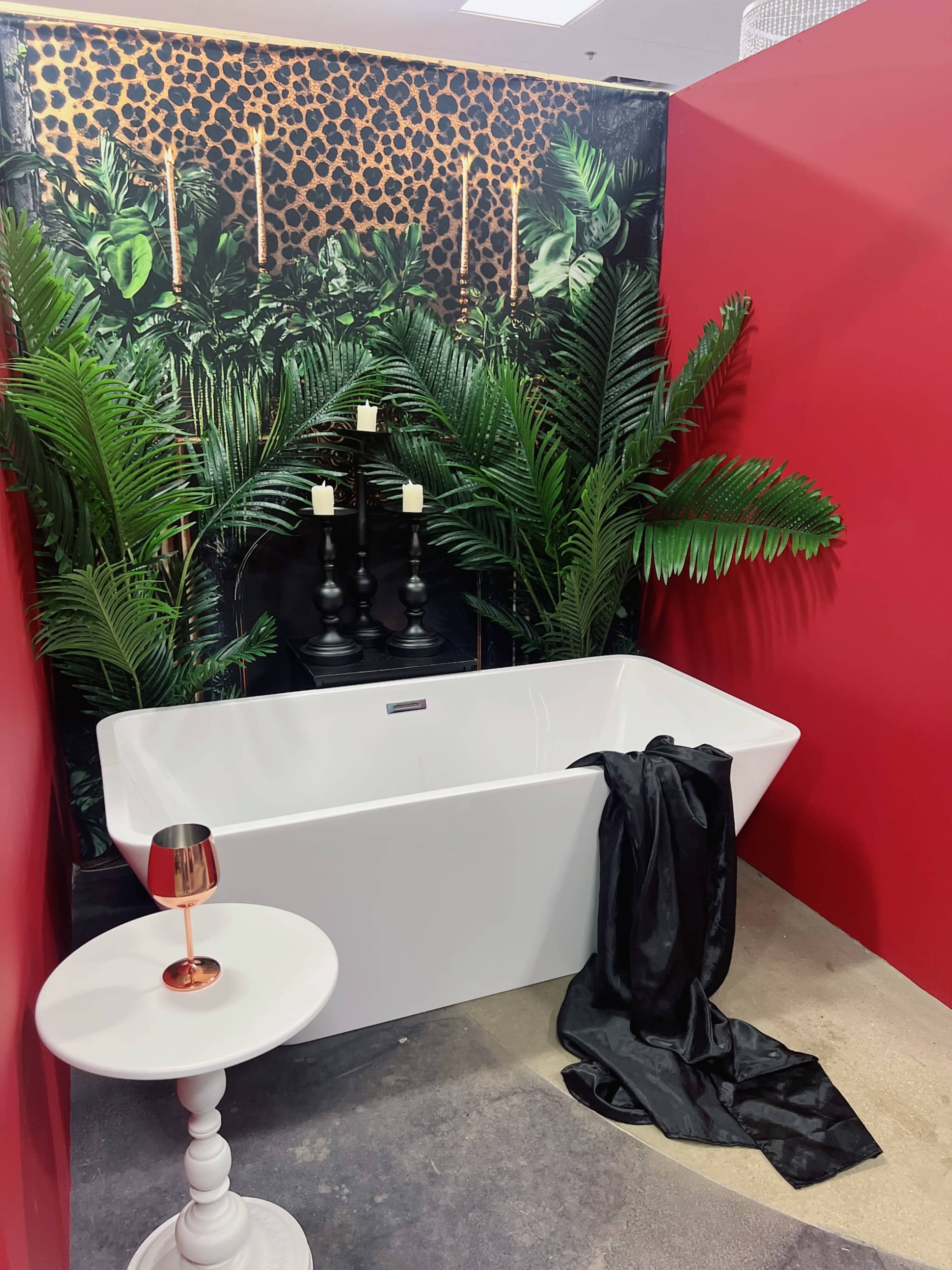 A modern white freestanding bathtub is positioned in front of a vibrant leopard print and foliage wallpaper, alongside a small white table with a copper-colored glass and a draped black fabric.