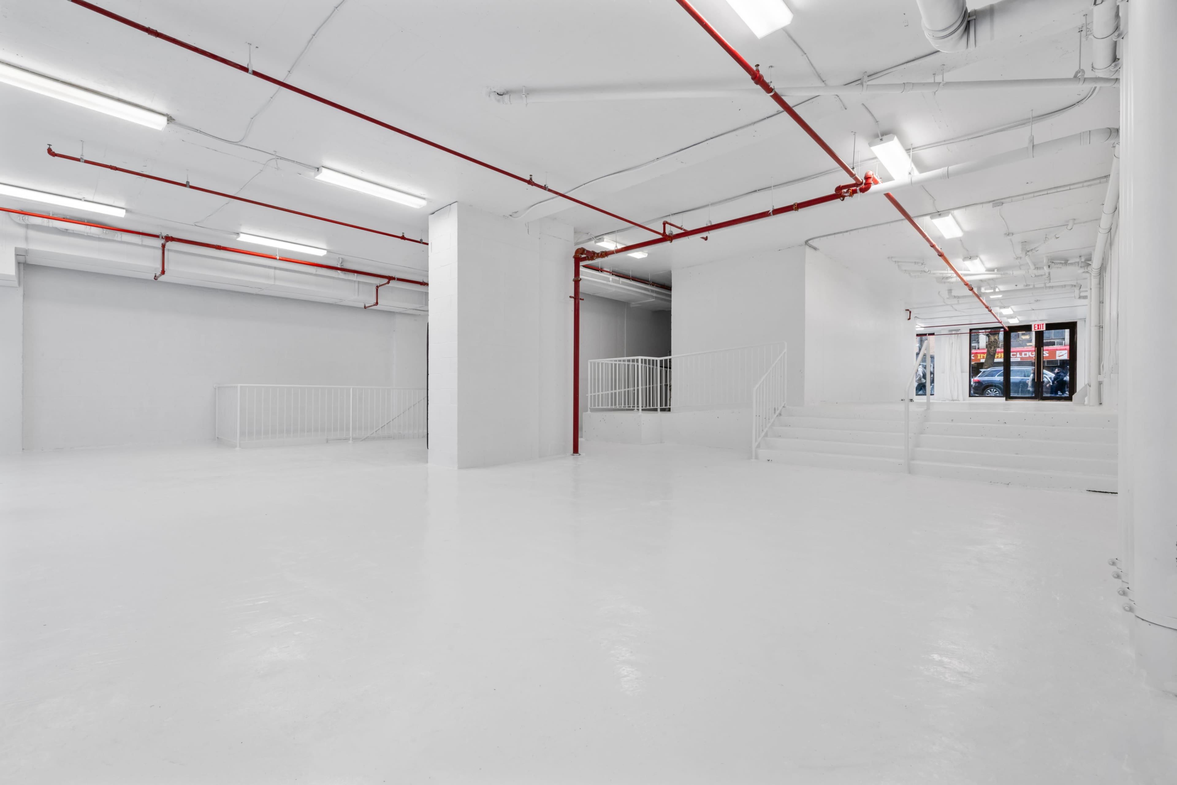 A large, empty commercial space with white walls and floors, featuring red piping along the ceiling and a staircase leading to an entrance.