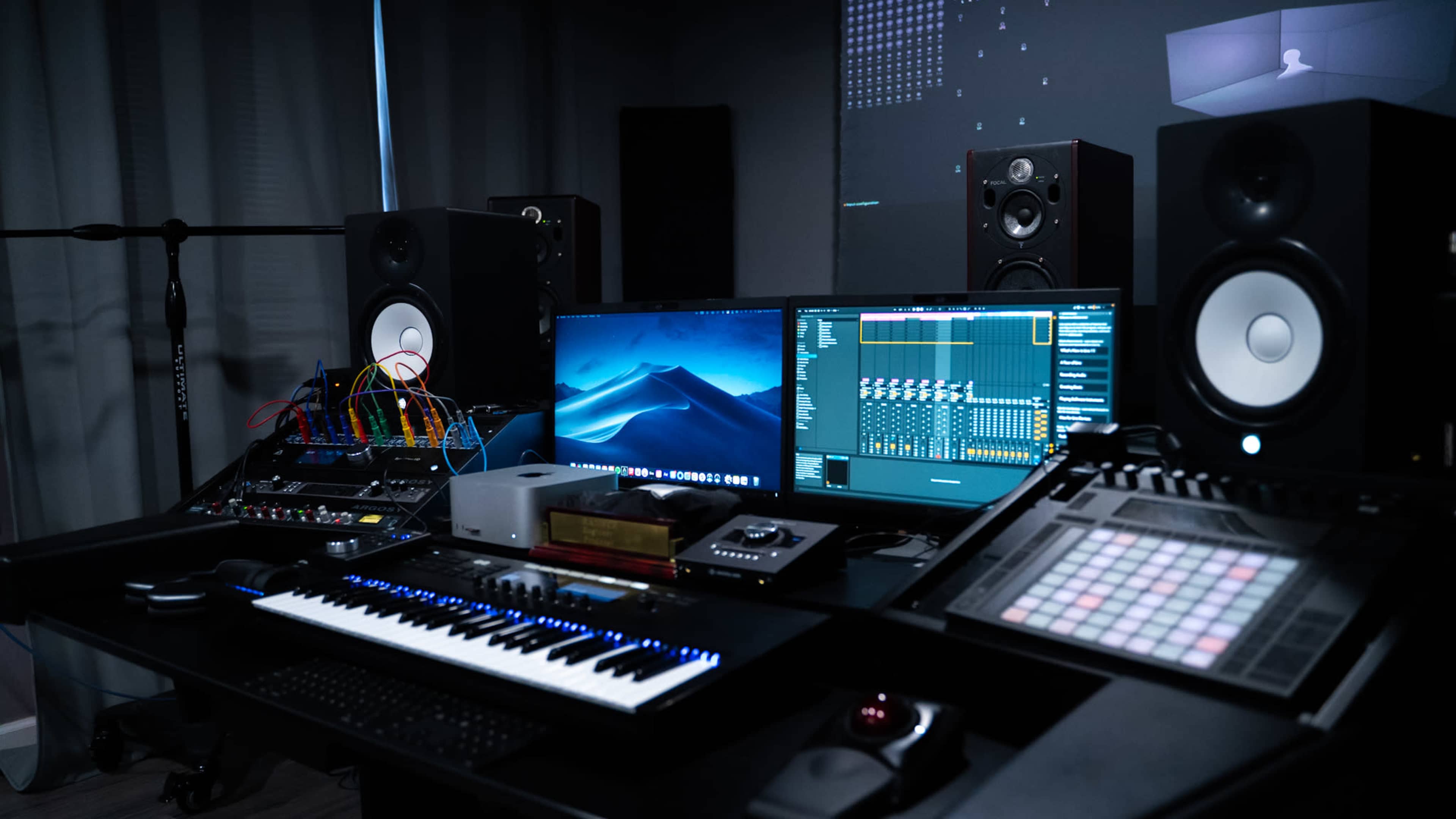 A modern music production studio features a keyboard, multiple monitors, audio equipment, and various sound modules arranged on a sleek desk.