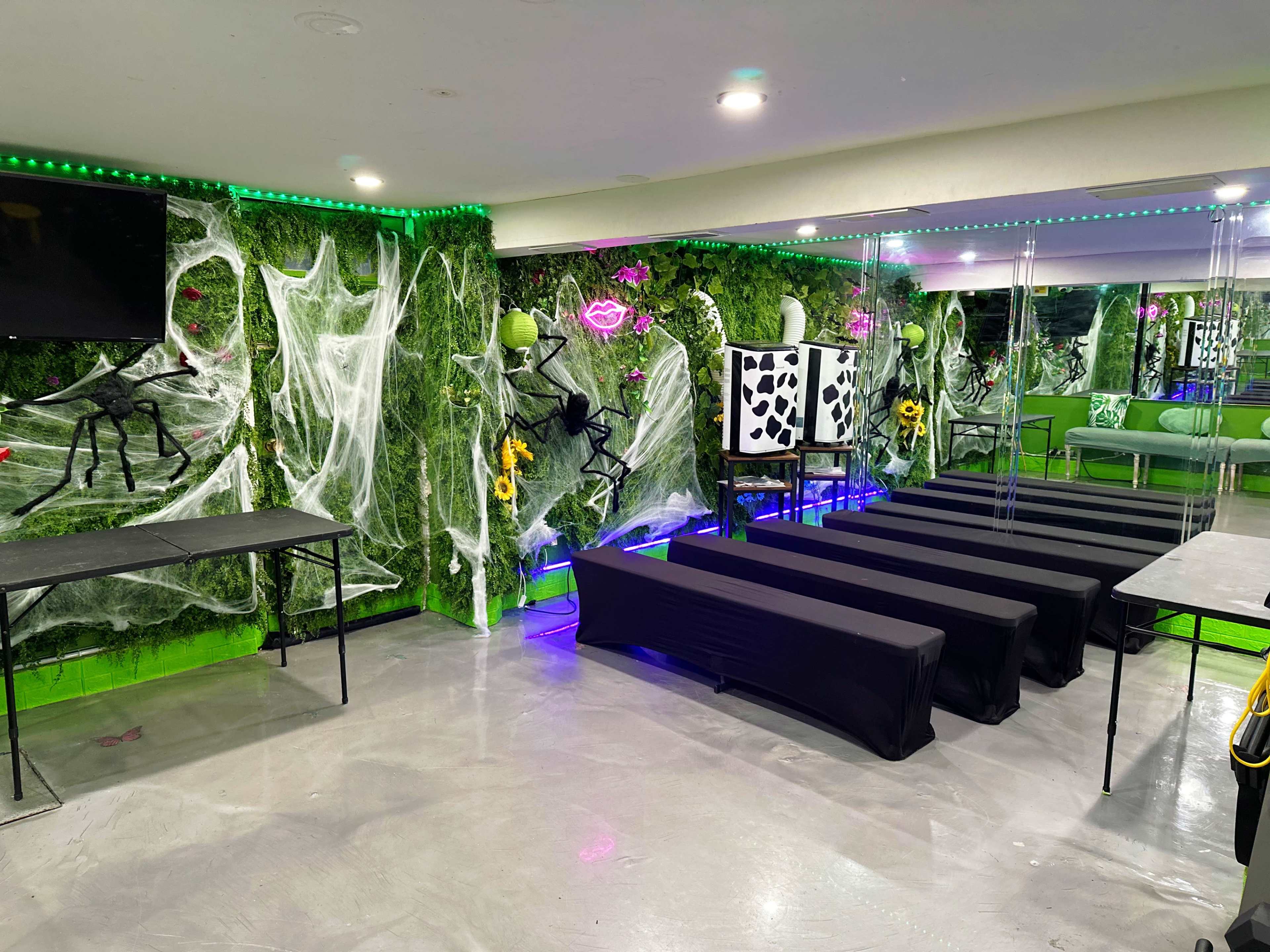 The room features a green wall decorated with artificial plants, spider webs, and neon lights, with several black tables arranged in a row.
