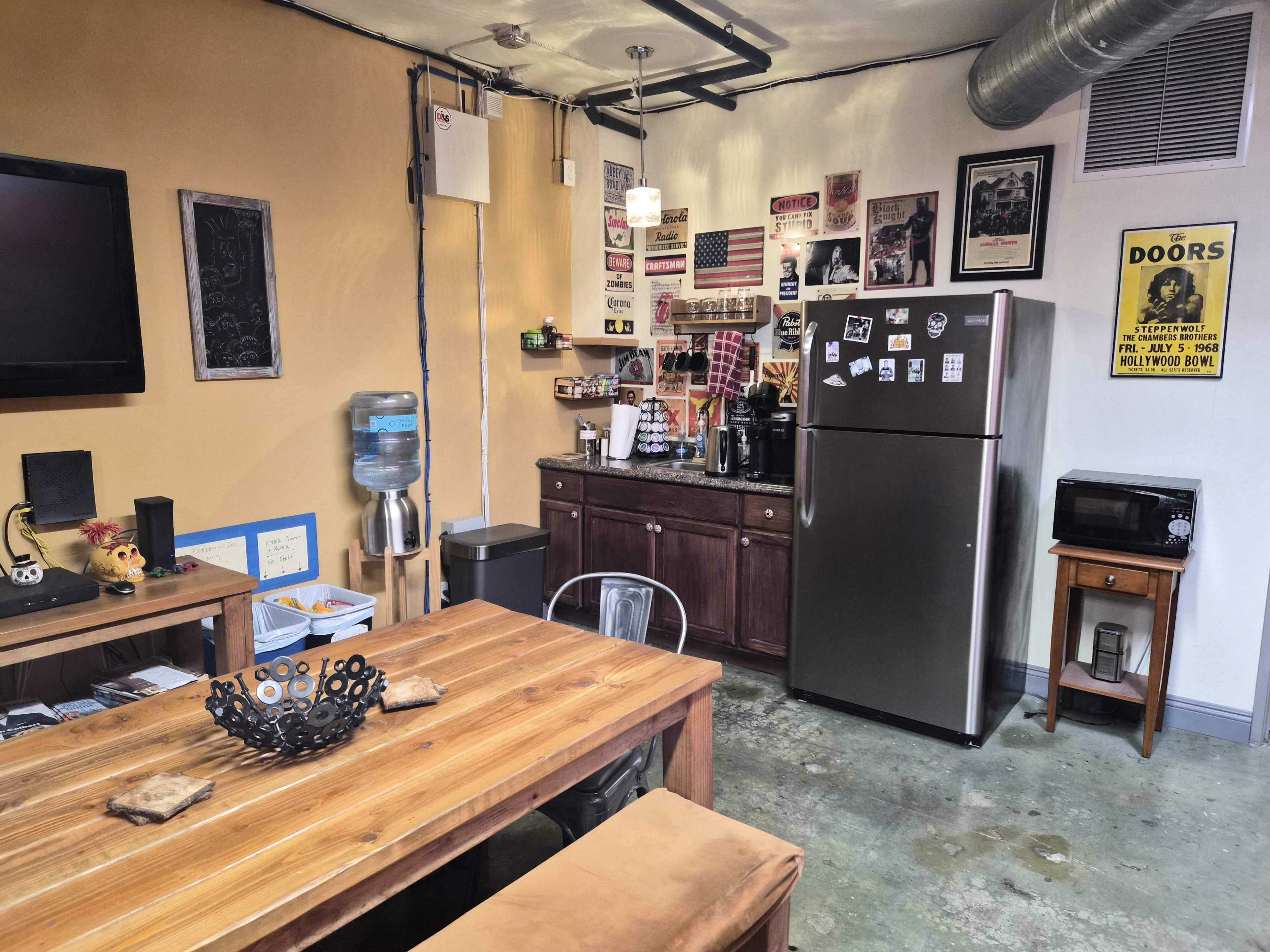 The image shows a kitchenette area featuring a stainless steel refrigerator, a water cooler, wooden dining table, and a variety of wall decorations.