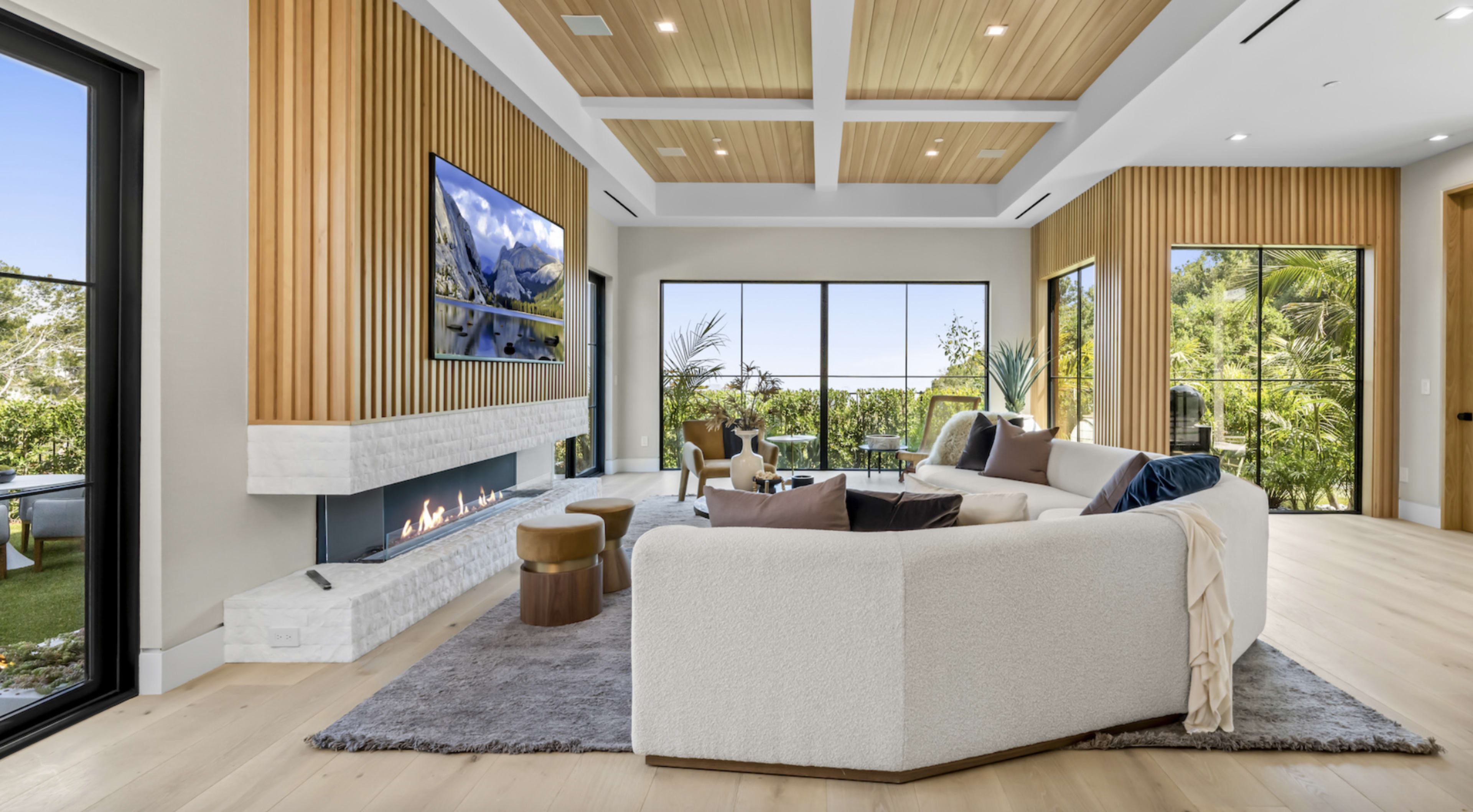 The image shows a modern living room featuring a large sectional sofa, a fireplace, a mounted television, and expansive windows offering a view of greenery outside.