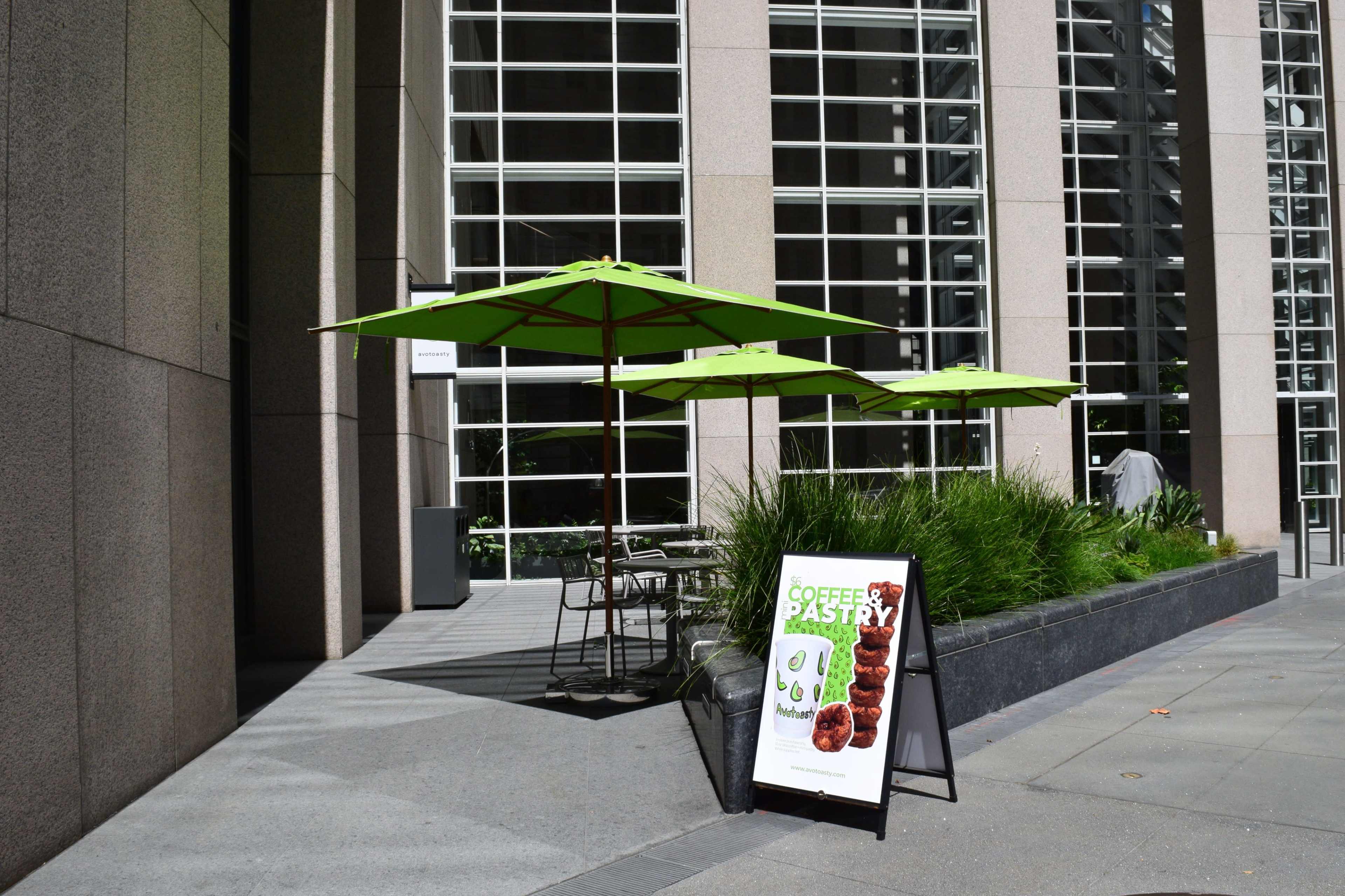 The image shows a sidewalk cafe with green umbrellas and a promotional sign for coffee and pastry in front of a modern building.