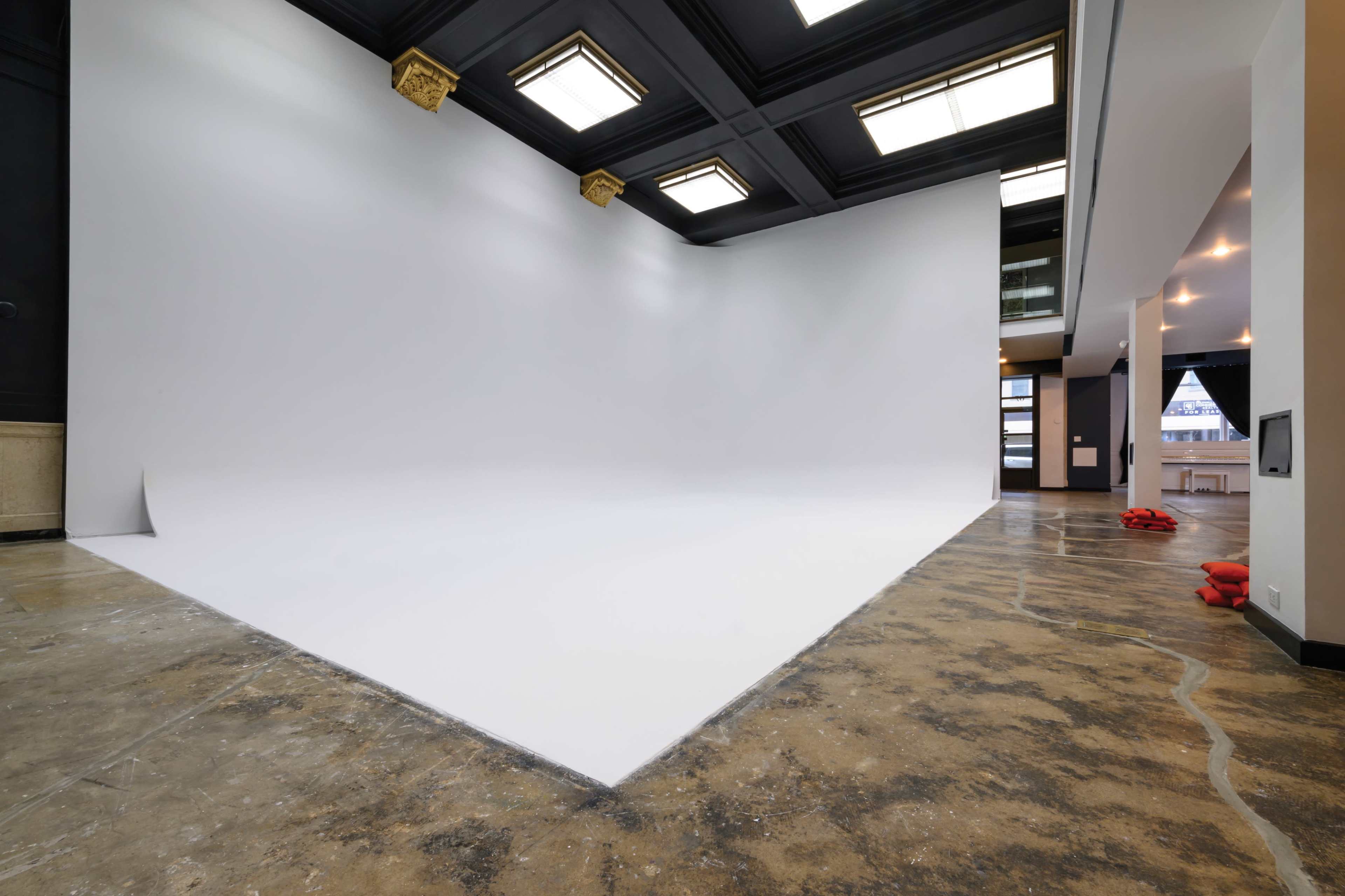 A spacious studio features a large, white backdrop against a polished concrete floor and black ceiling, with soft lighting and minimal furnishings.