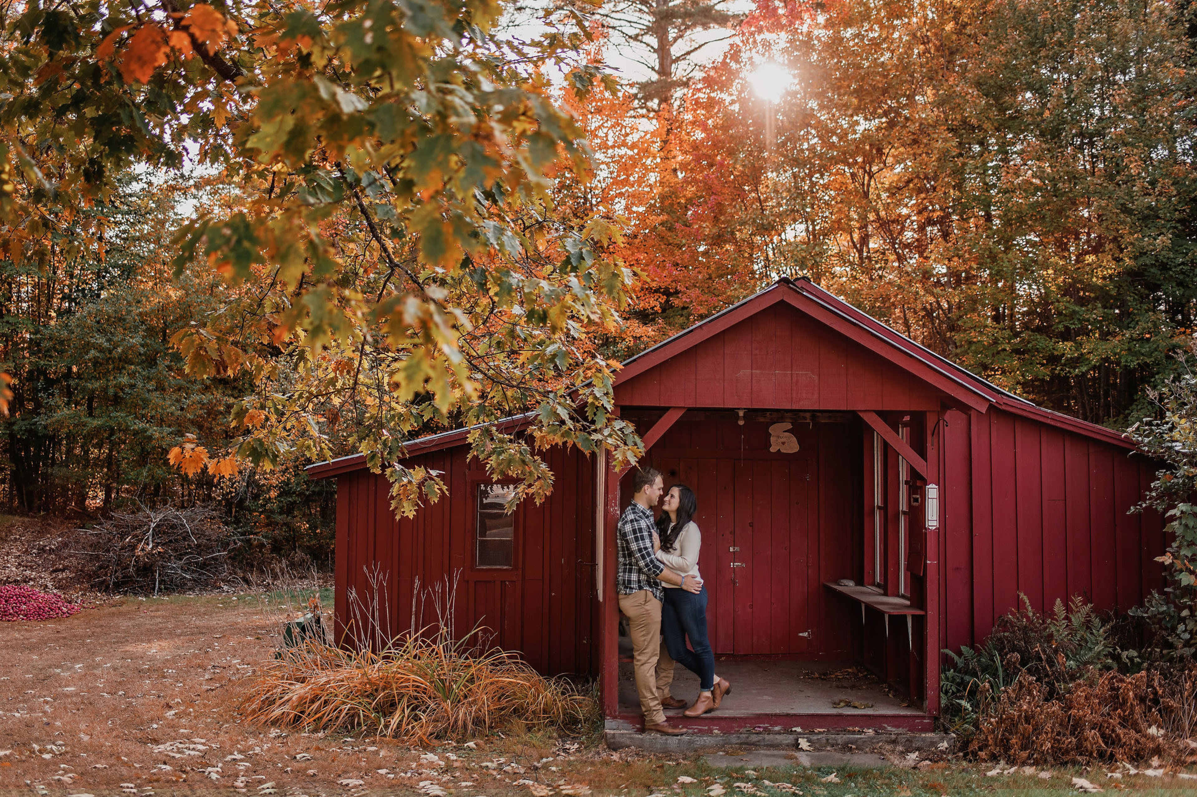 A couple stands outside a red cabin surrounded by autumn-colored trees.