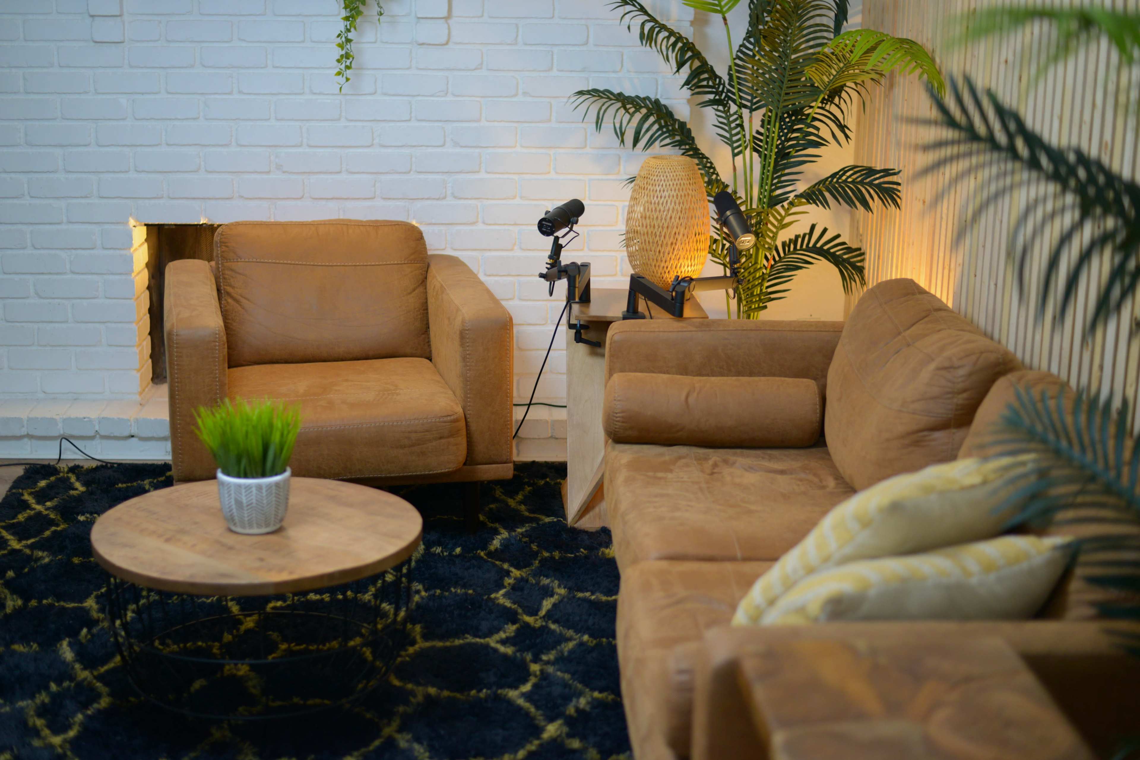 A cozy living room features two brown leather chairs, a round wooden coffee table, and a potted green plant, set against a white brick wall.