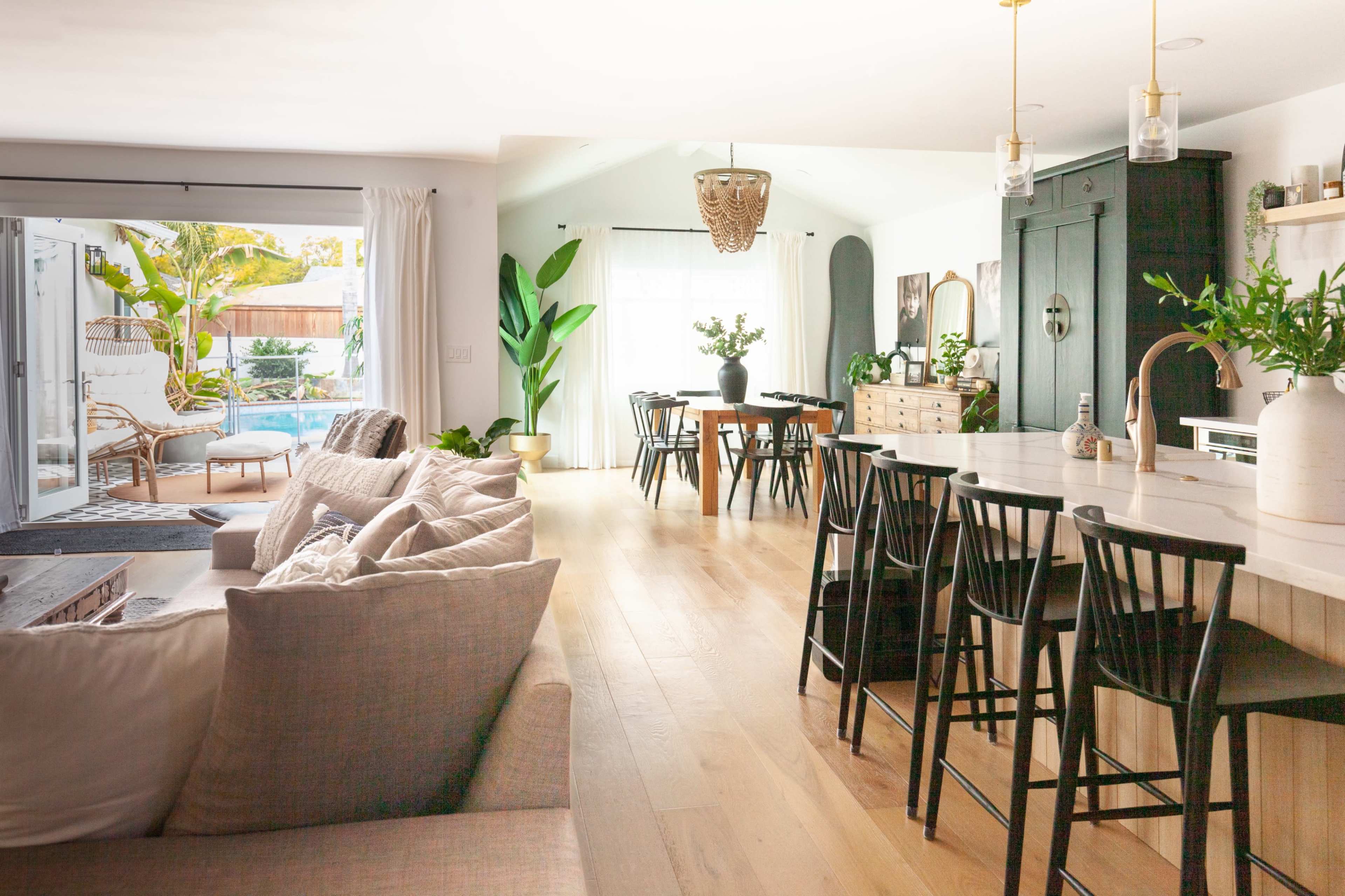 A bright and open living space features a kitchen island with bar stools, a dining table, and a lounge area with a sectional sofa, leading to a pool area visible through sliding glass doors.