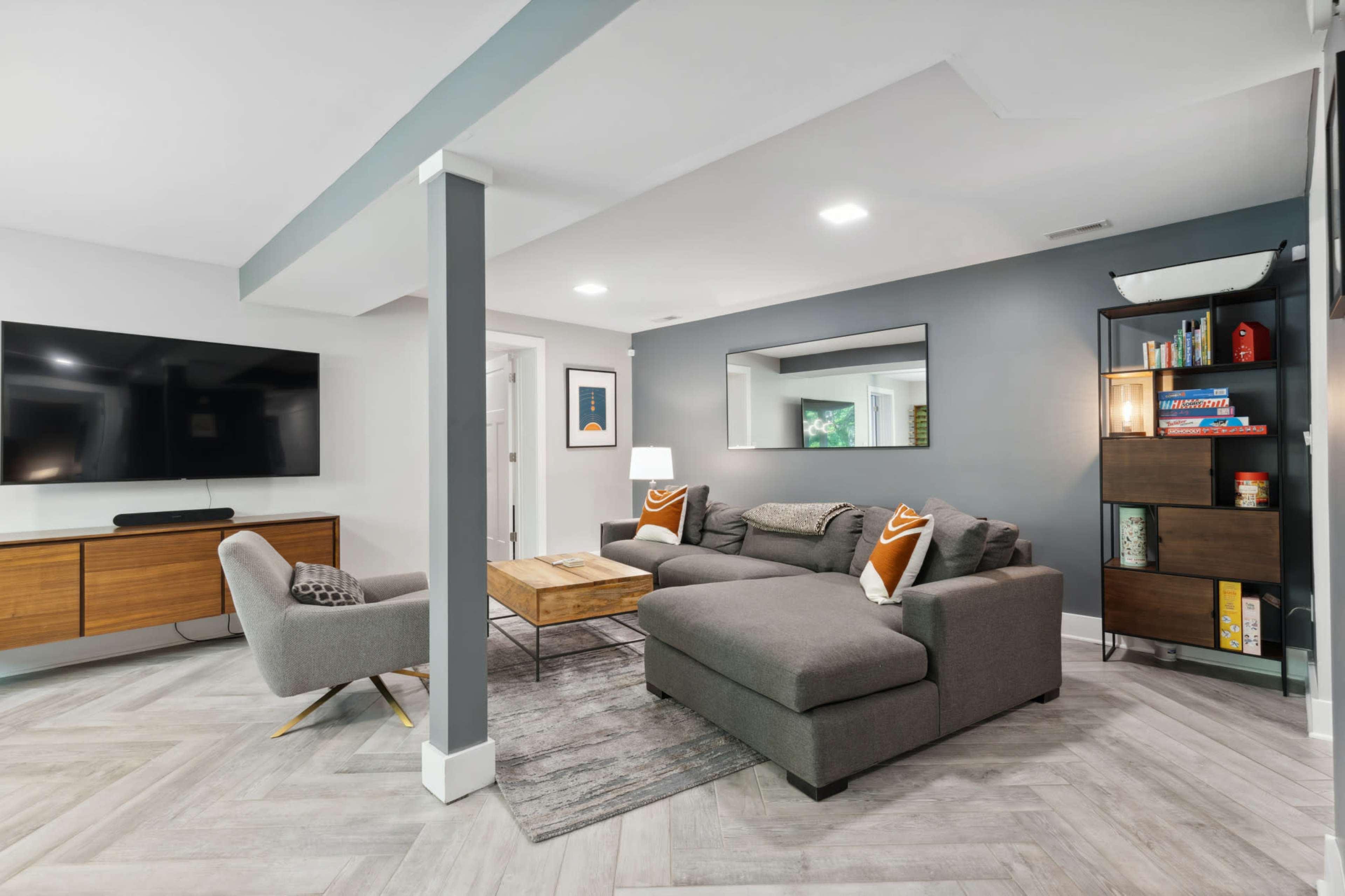 The image shows a modern living room with a gray sectional sofa, a wooden coffee table, a television mounted on the wall, and a bookshelf, all set against a light-colored wall with decorative accents.