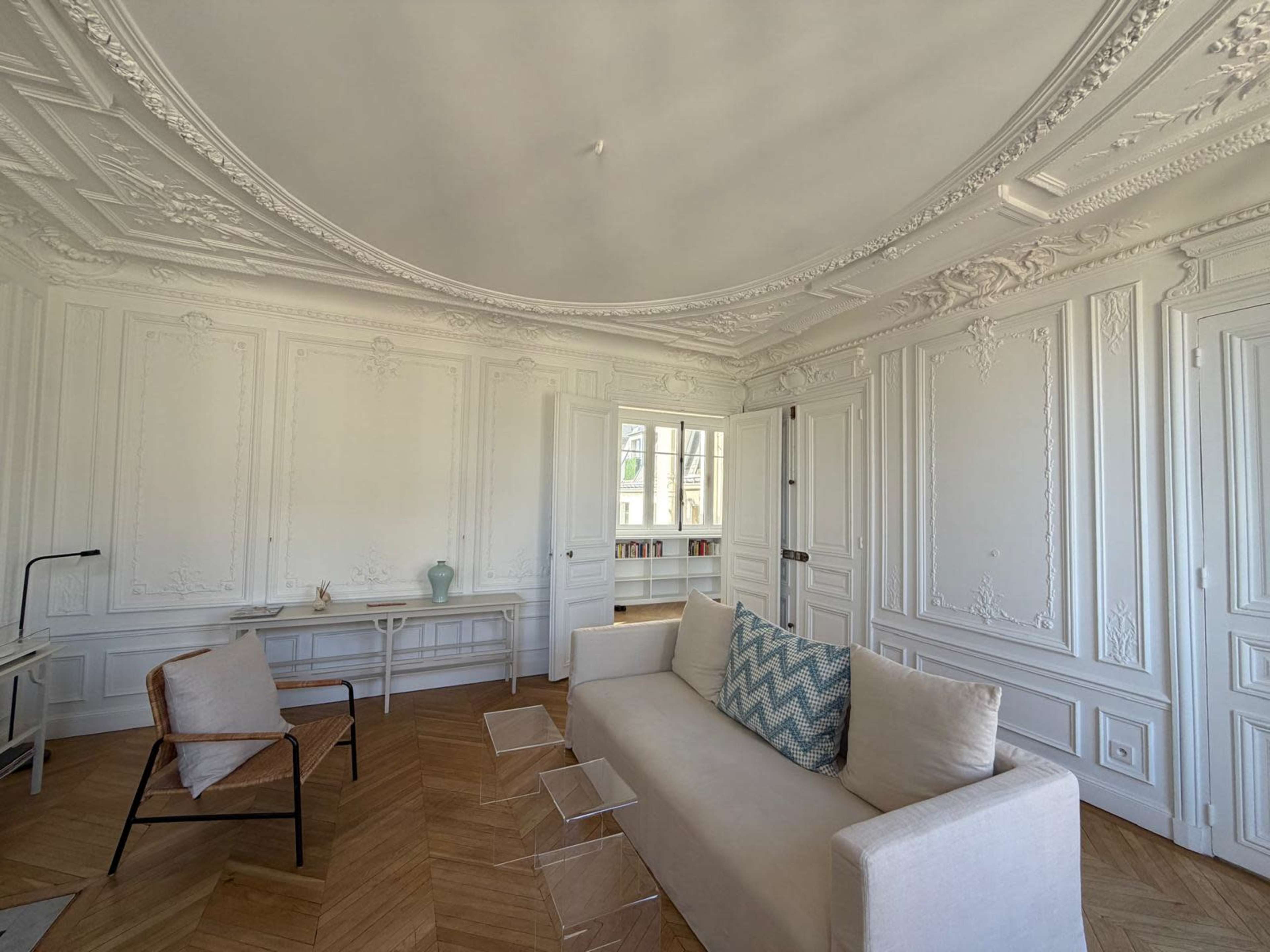 The image shows a bright, elegantly designed living room featuring a white sofa with a decorative pillow, a small coffee table, and ornate wall moldings, complemented by natural light coming through large windows.