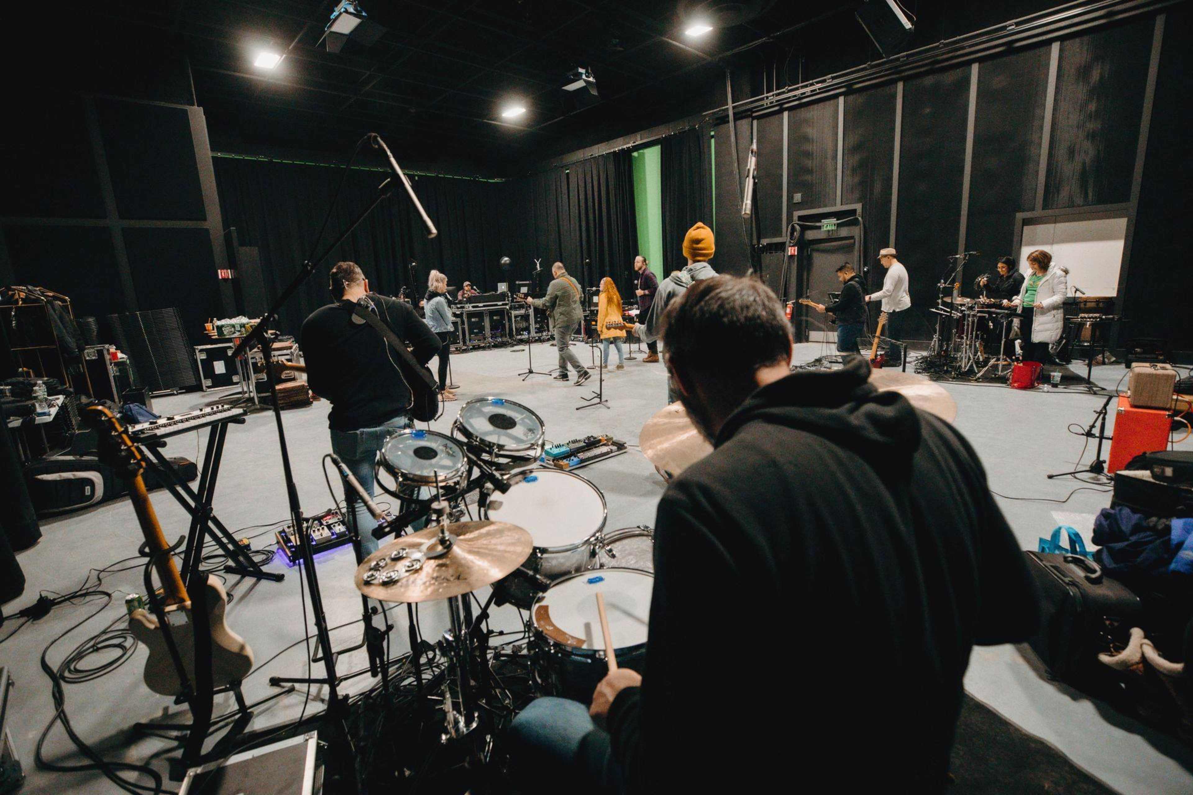 A group of musicians rehearses in a spacious studio with various instruments set up, including a drum kit in the foreground.