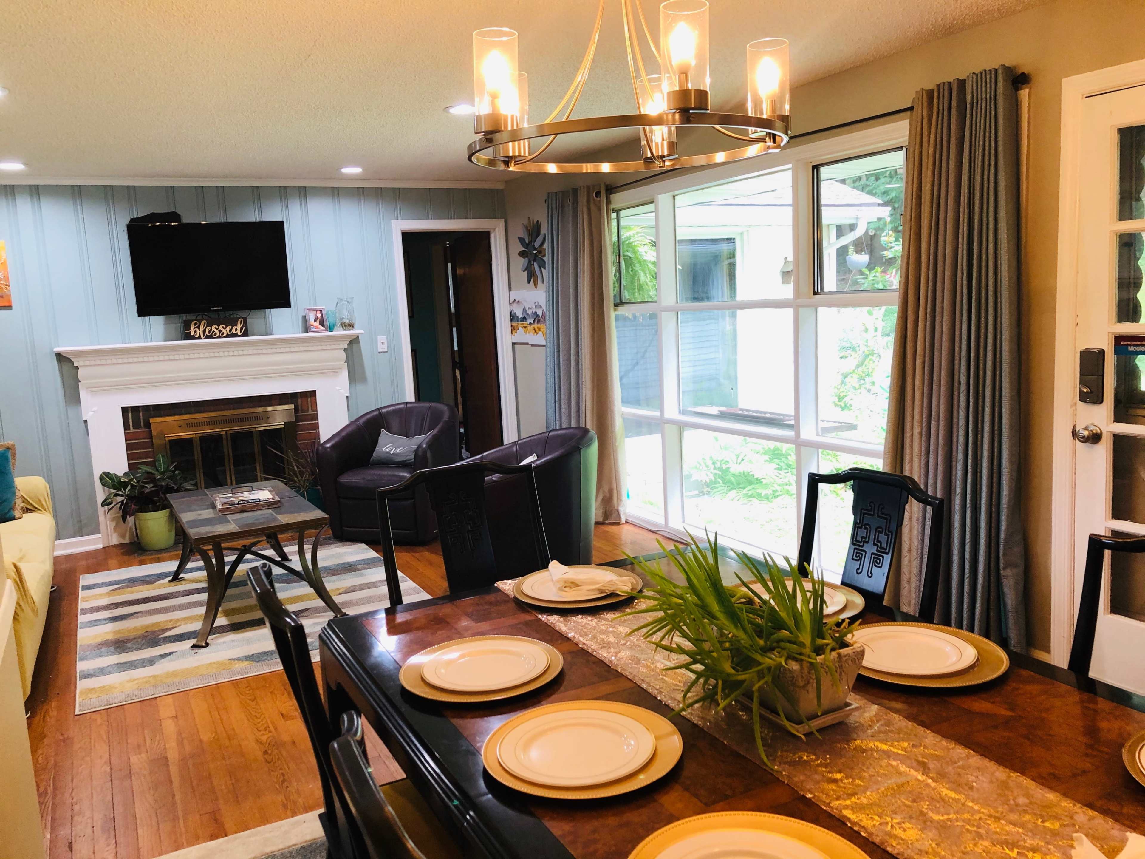 A dining area with a wooden table set for a meal, surrounded by chairs, adjacent to a living space featuring a fireplace and a large window overlooking a garden.