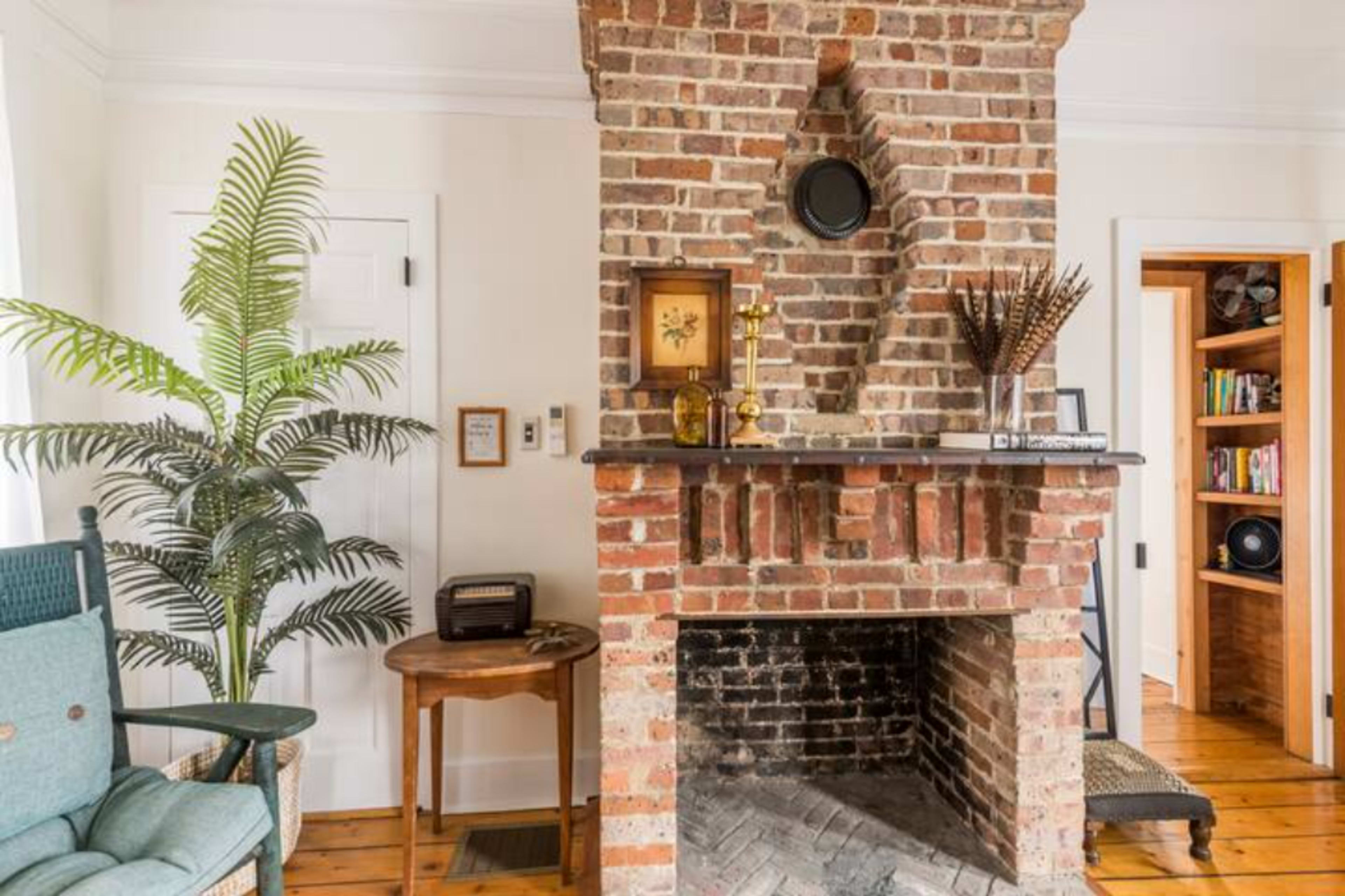 The image shows a brick fireplace with a wooden mantel, surrounded by a plant, a small table, and a door leading to a bookshelf.