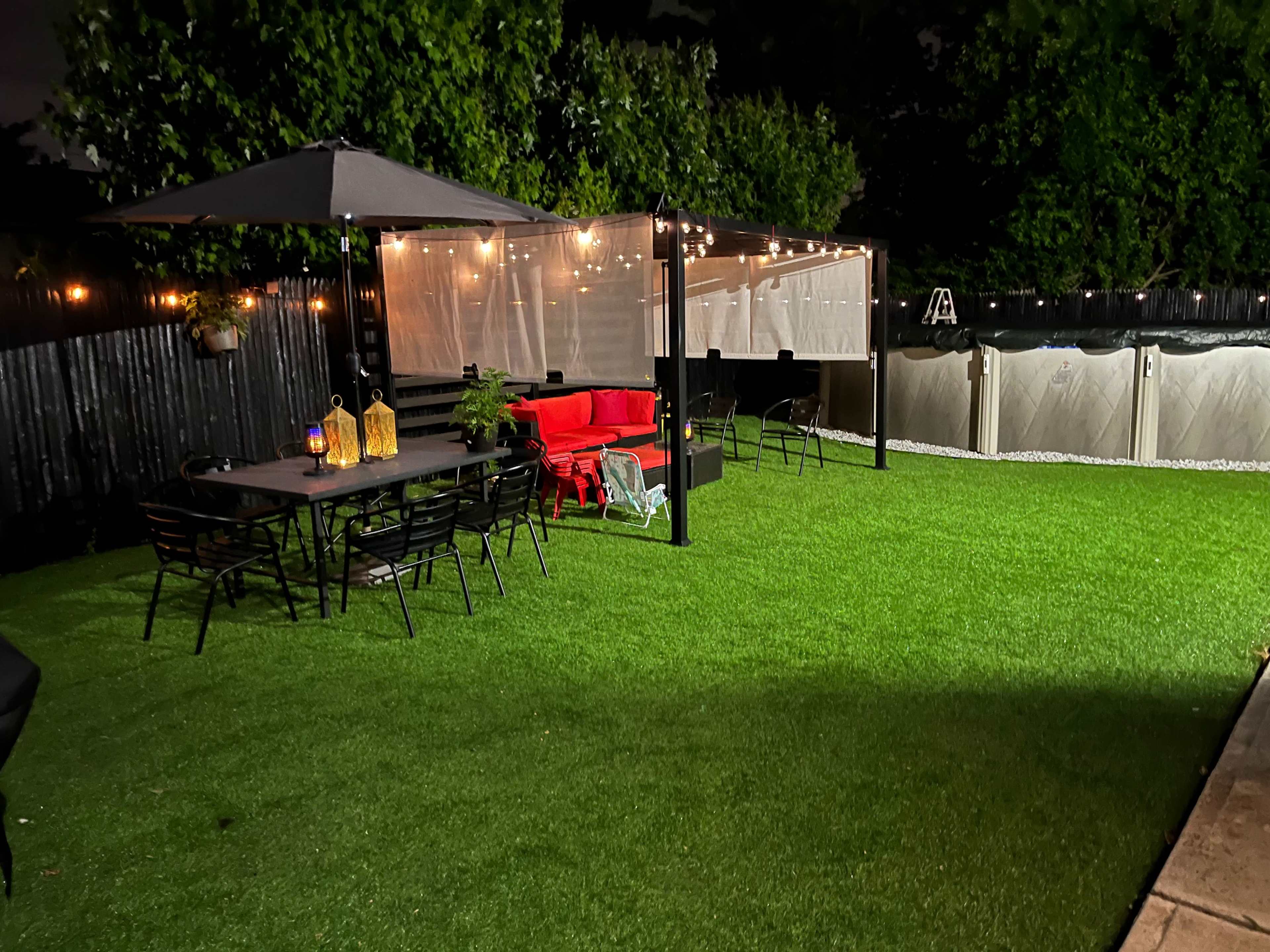 A well-lit backyard with an artificial grass lawn, a dining table with chairs, a covered seating area with a red couch, and string lights overhead.