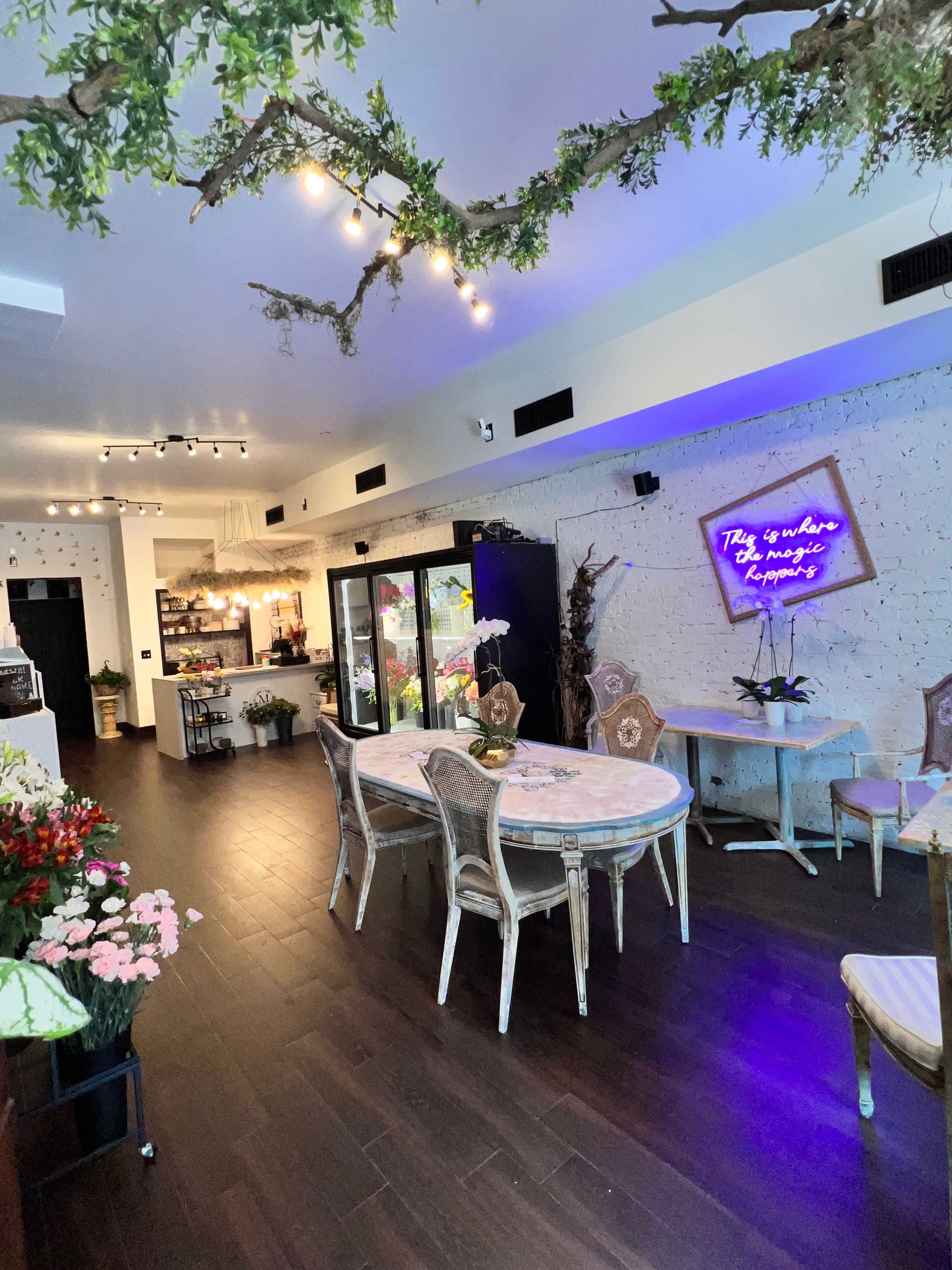 A modern floral shop with a bright interior, featuring a dining table surrounded by chairs, a display case filled with flowers, and a neon sign on the wall.