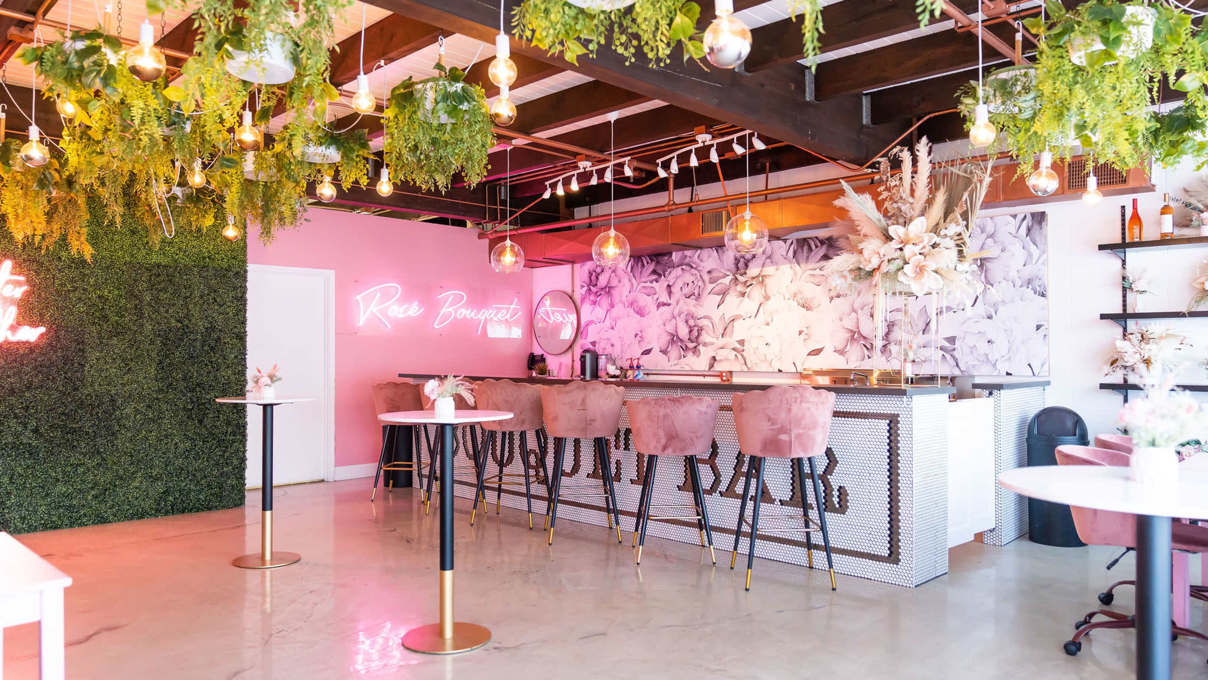 A brightly adorned bar area with pink seating, greenery hanging from the ceiling, and a floral mural on the wall.