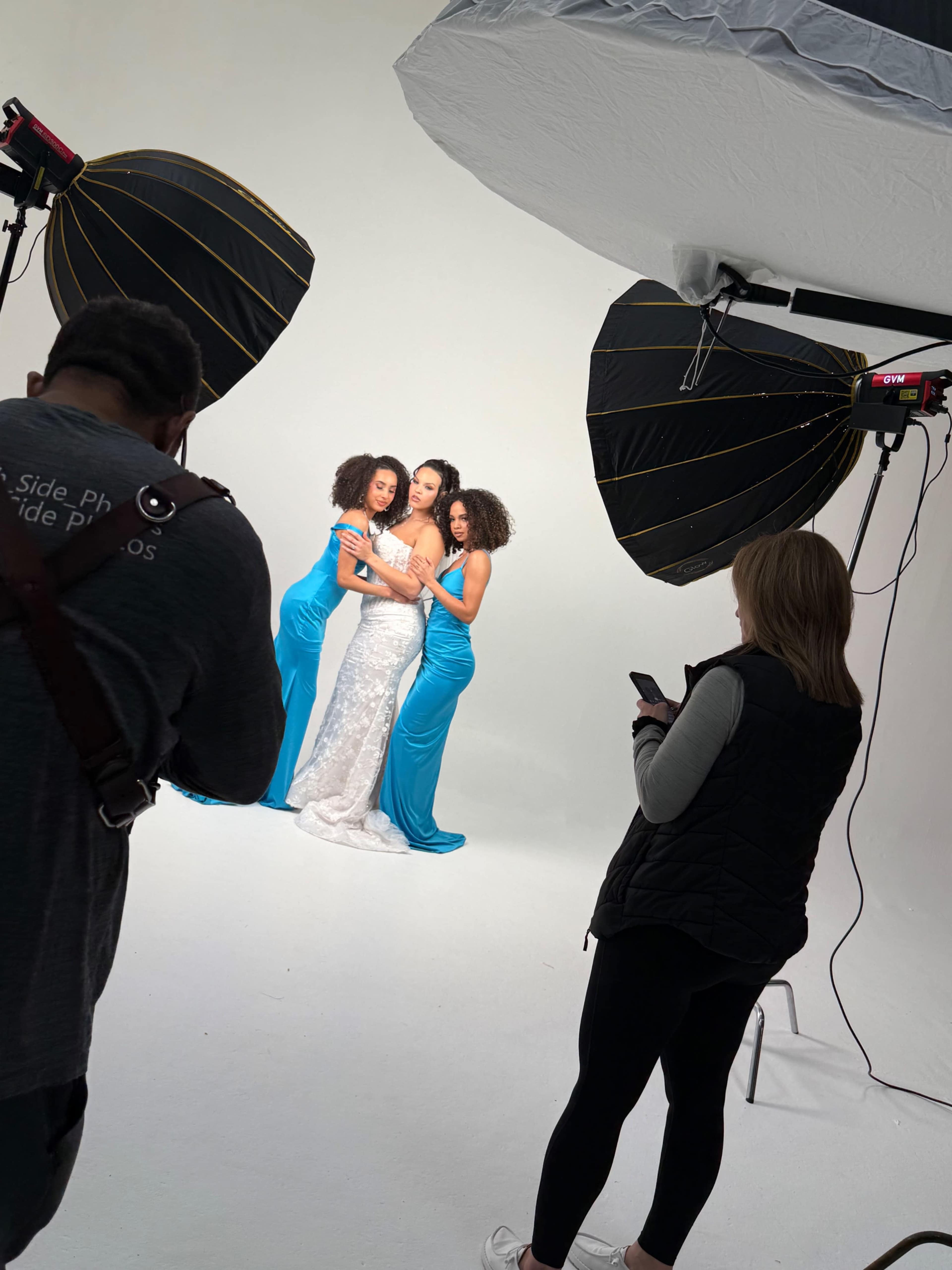 A photographer captures three women posing together in elegant outfits against a plain white backdrop, while another person checks their phone nearby.